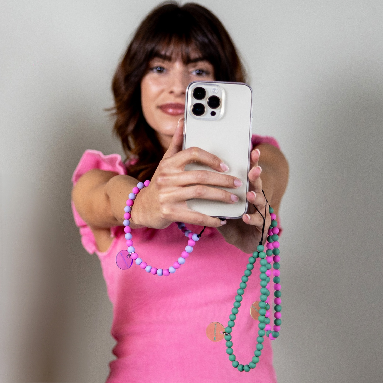Donna in rosa tiene in mano un telefono con cinturini di perline. Le perline sono rosa, viola e verdi. Lo sfondo è grigio chiaro.