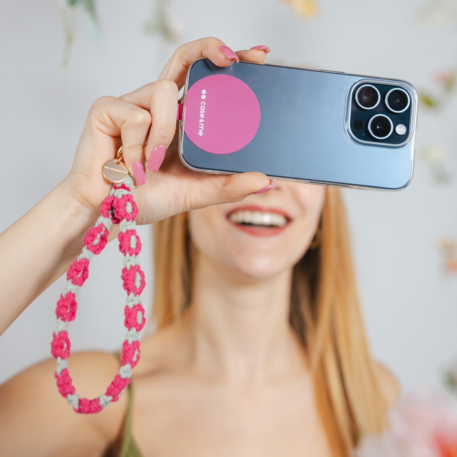 Donna che tiene in mano telefono con cerchio rosa, cinturino abbinato. Unghie rosa. Sorridente. Sfondo chiaro con accenti floreali.