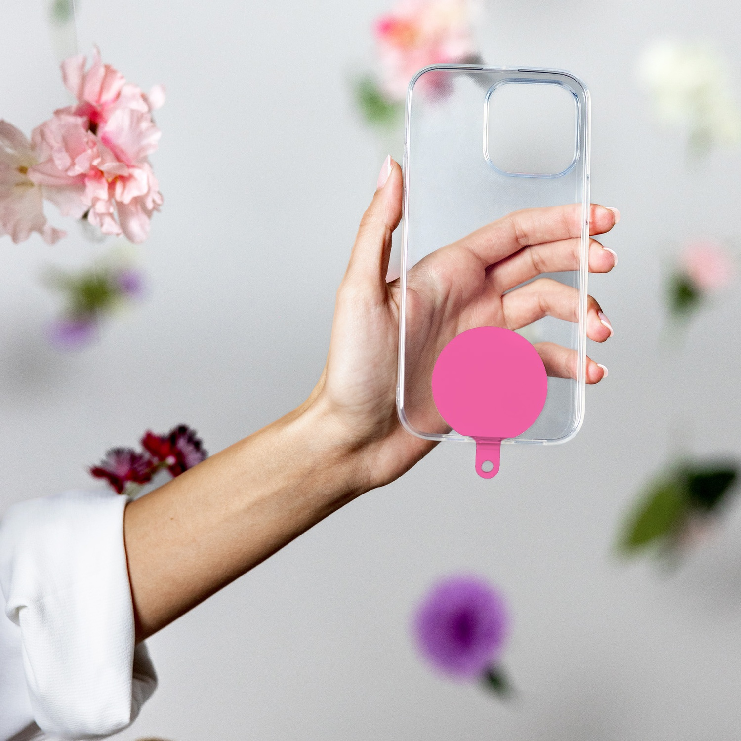 Una mano tiene una custodia trasparente per telefono con un cerchio rosa. Fiori appesi sullo sfondo, contro un muro grigio chiaro.