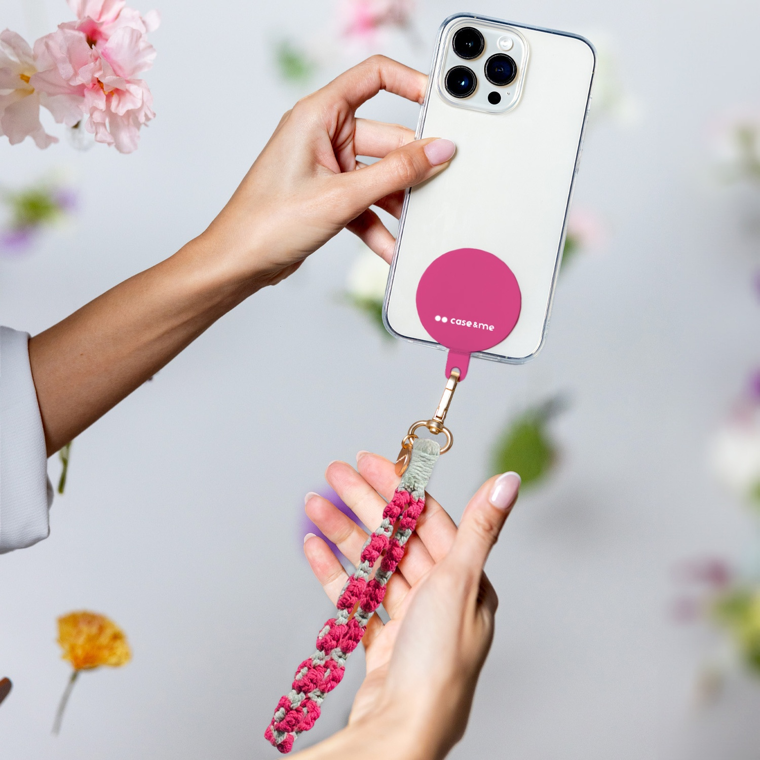 Mani che tengono un telefono con custodia rosa e cinturino in macramè, circondato da fiori. Il telefono è bianco con un cerchio rosa.