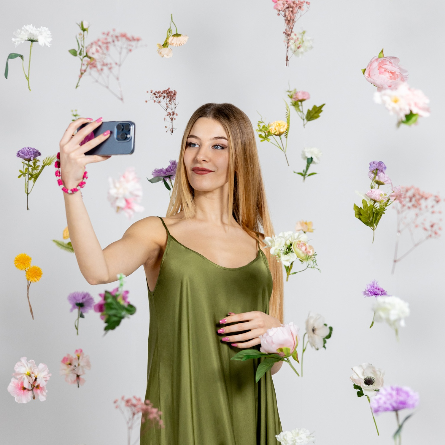 Donna in abito verde fa un selfie circondata da fiori fluttuanti. Braccialetto e unghie rosa. Sfondo bianco.