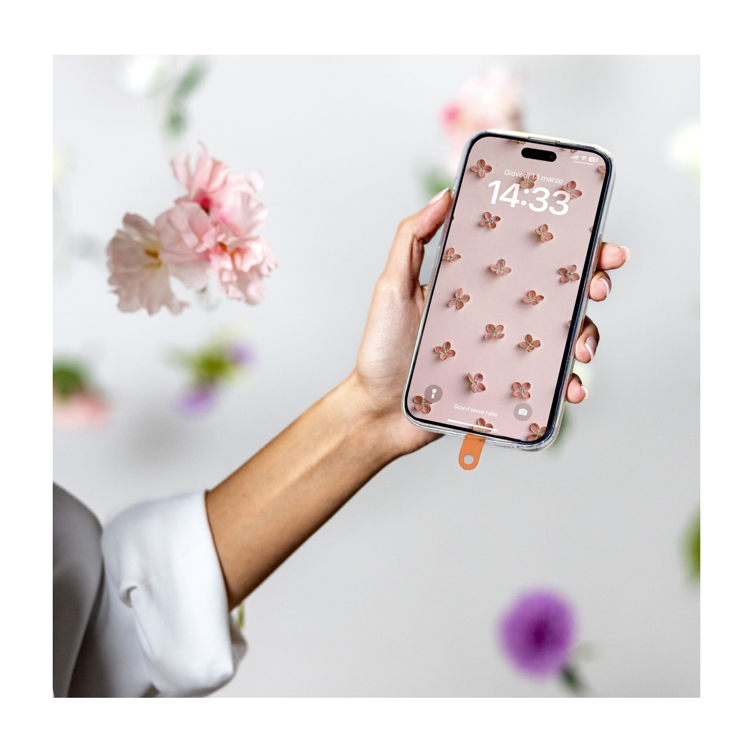 Mano che tiene il telefono con sfondo floreale. Fiori rosa fluttuano sullo sfondo. Manica bianca della camicia visibile.