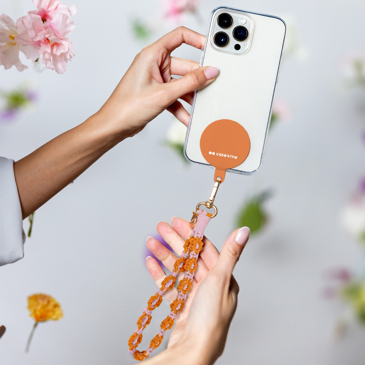 Mani che tengono un telefono con un'impugnatura arancione e un cinturino in macramè. Fiori sullo sfondo sfocato.