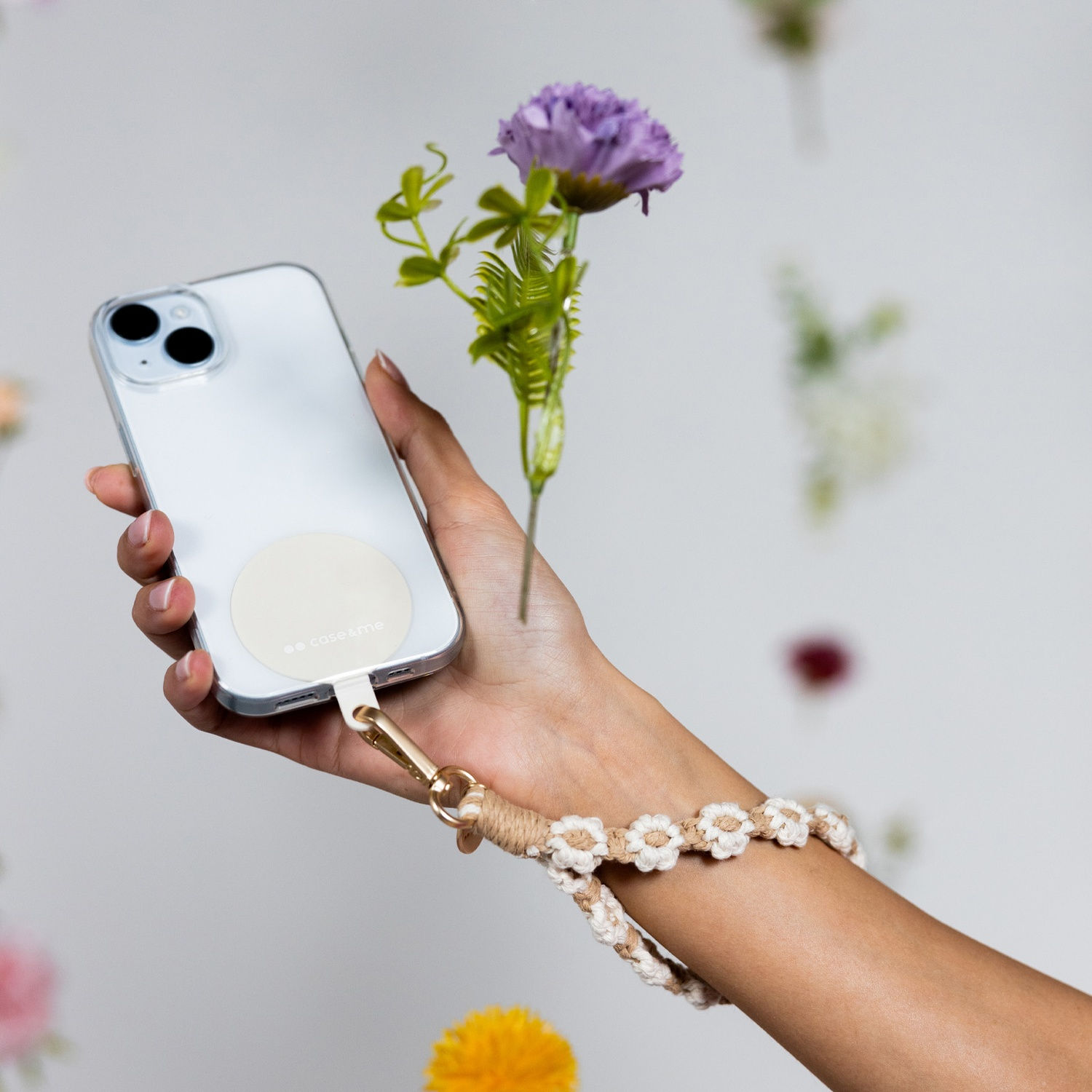 Mano che tiene un telefono con cinturino e fiori. Il telefono è bianco, il cinturino beige e i fiori viola.