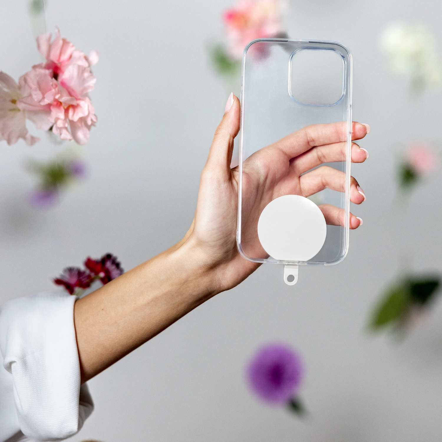 Una mano che tiene una custodia trasparente per telefono con un cerchio bianco, fiori sullo sfondo. La mano indossa una camicia bianca.