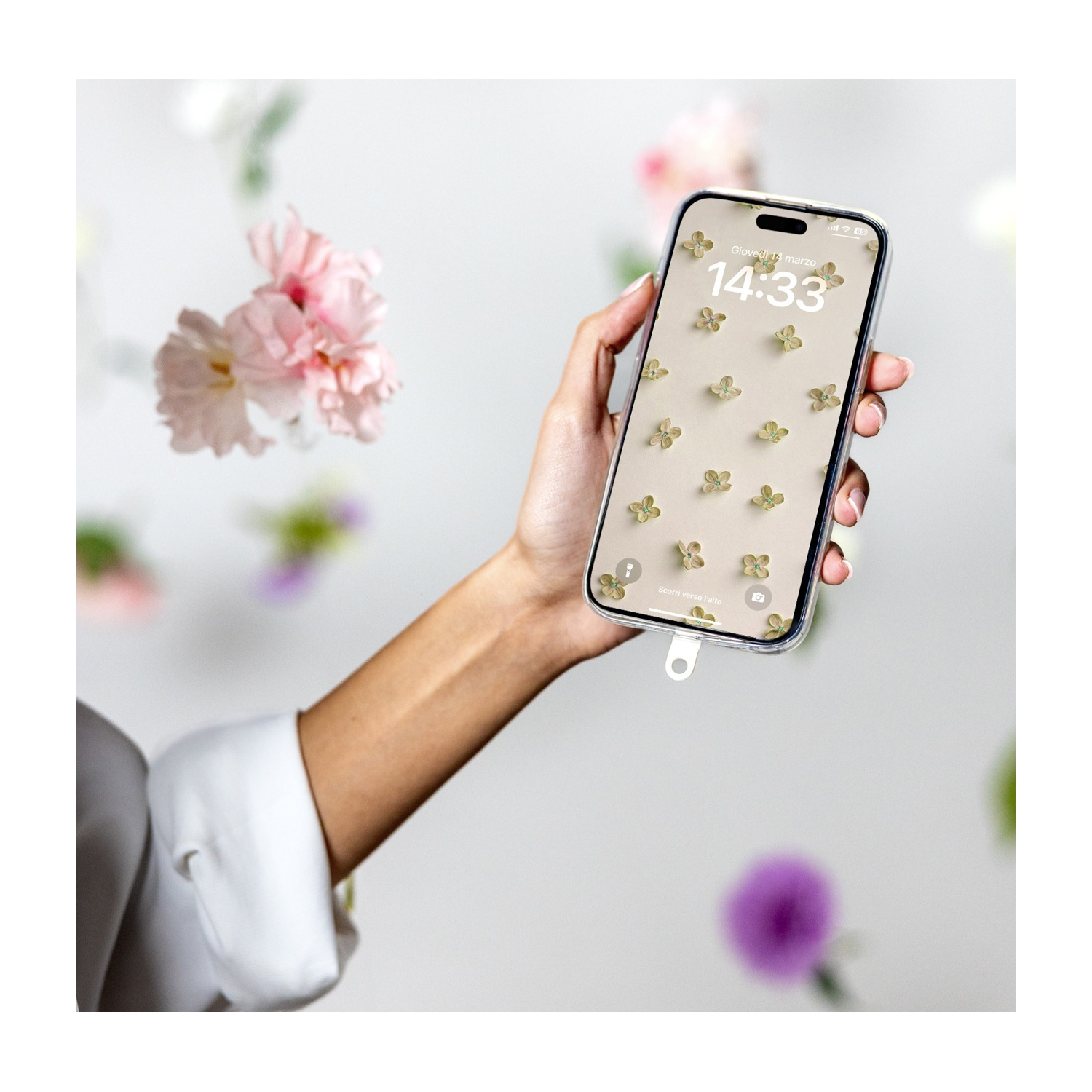 Una mano tiene un telefono con uno sfondo floreale. Fiori rosa fluttuano sullo sfondo. Il telefono mostra l'ora 14:33.