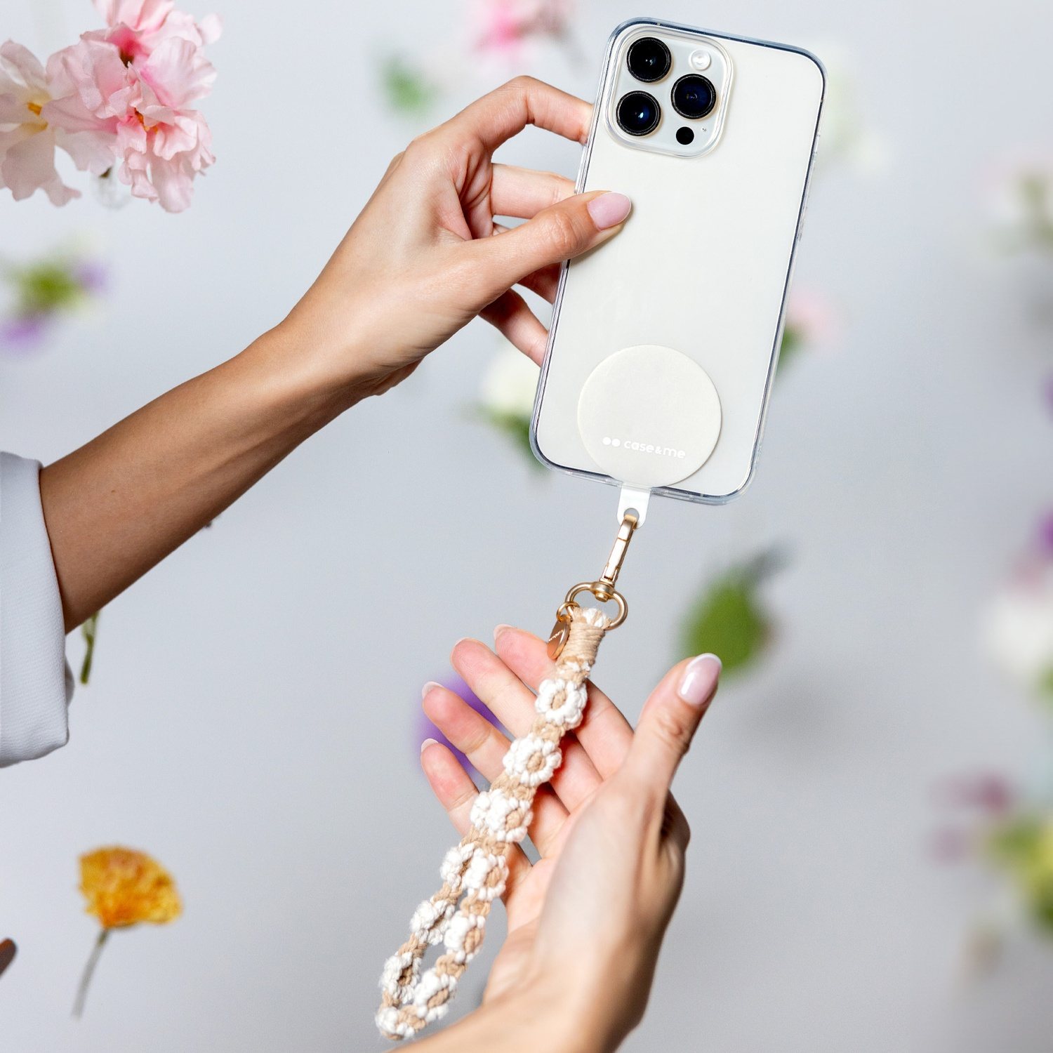 Mani che tengono un telefono con un cinturino. Il telefono è in una custodia trasparente. Fiori sullo sfondo.