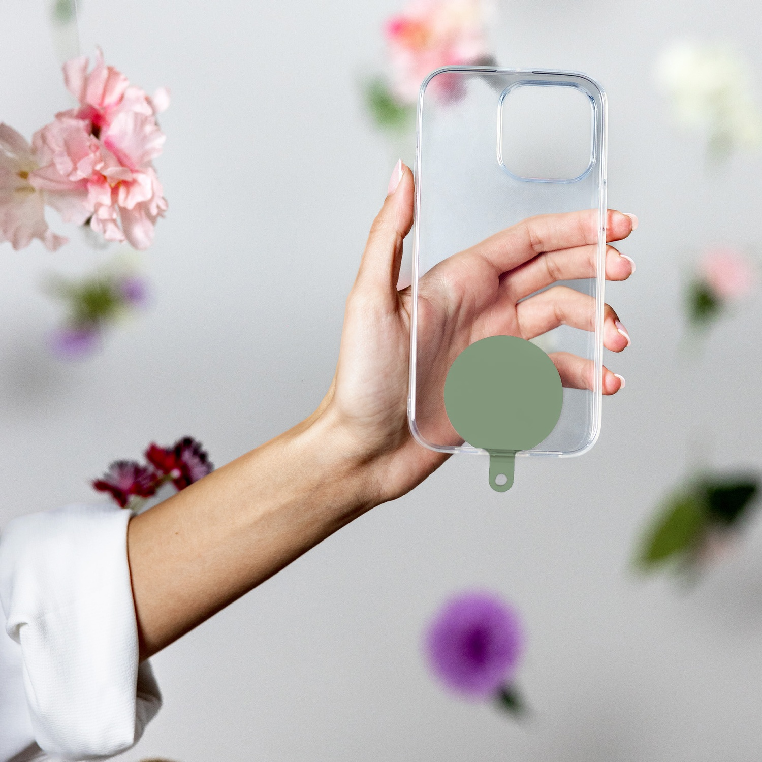 Mano che tiene una custodia trasparente con impugnatura circolare verde, fiori sullo sfondo. Manica di camicia bianca visibile.