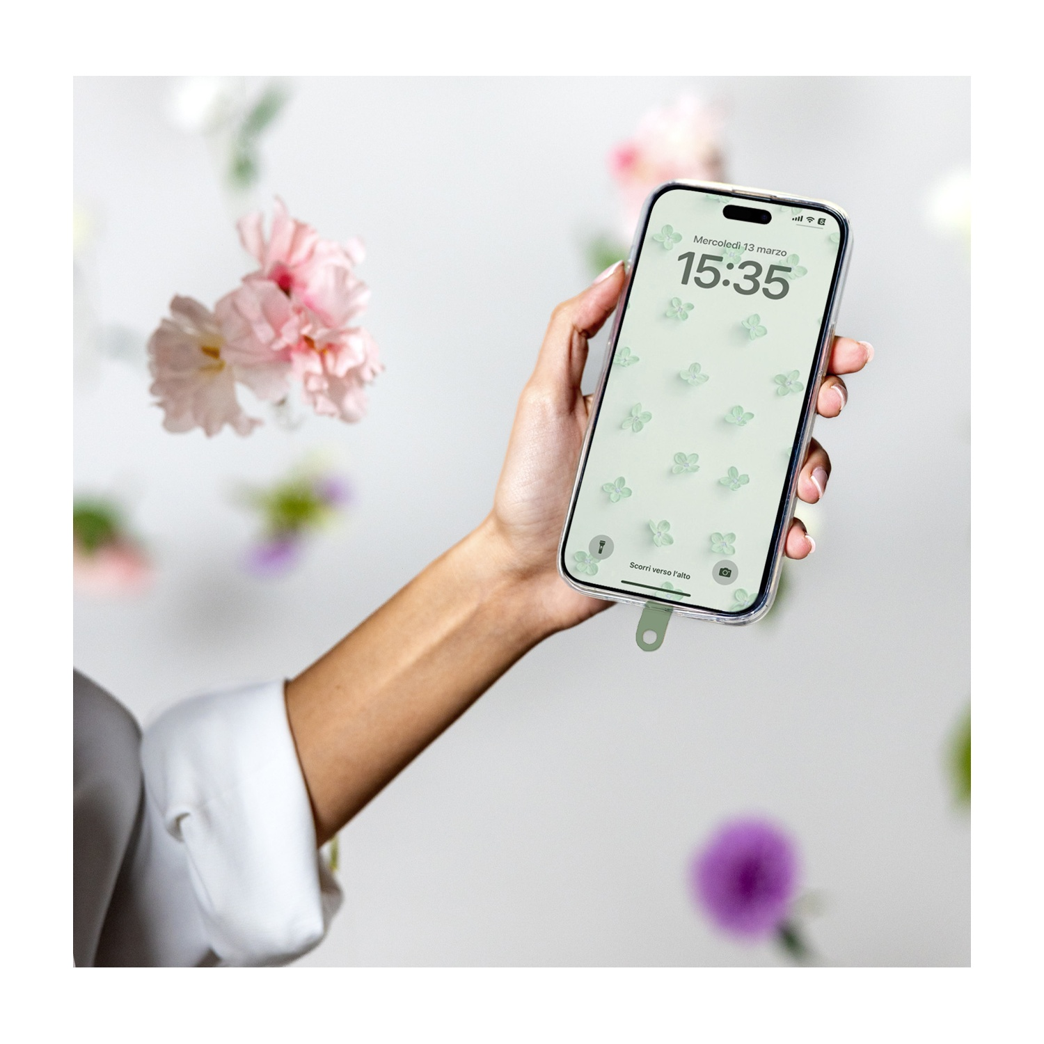Una mano tiene un telefono con uno sfondo floreale. Fiori rosa fluttuano nello sfondo sfocato. L'ora è 15:35.