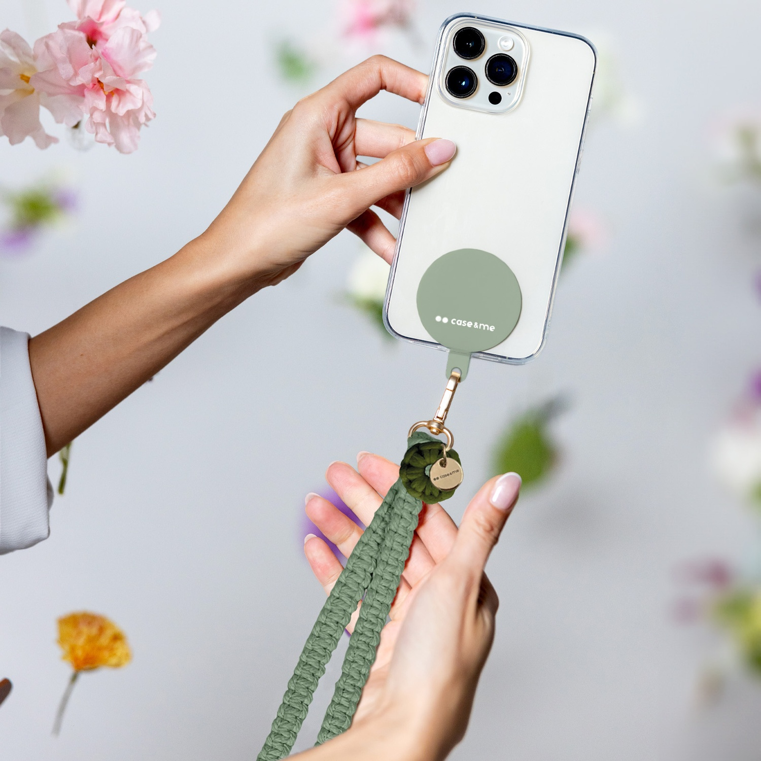 Mani che tengono un telefono con cinturino. Il telefono ha una custodia e un cinturino verde. Fiori sullo sfondo.