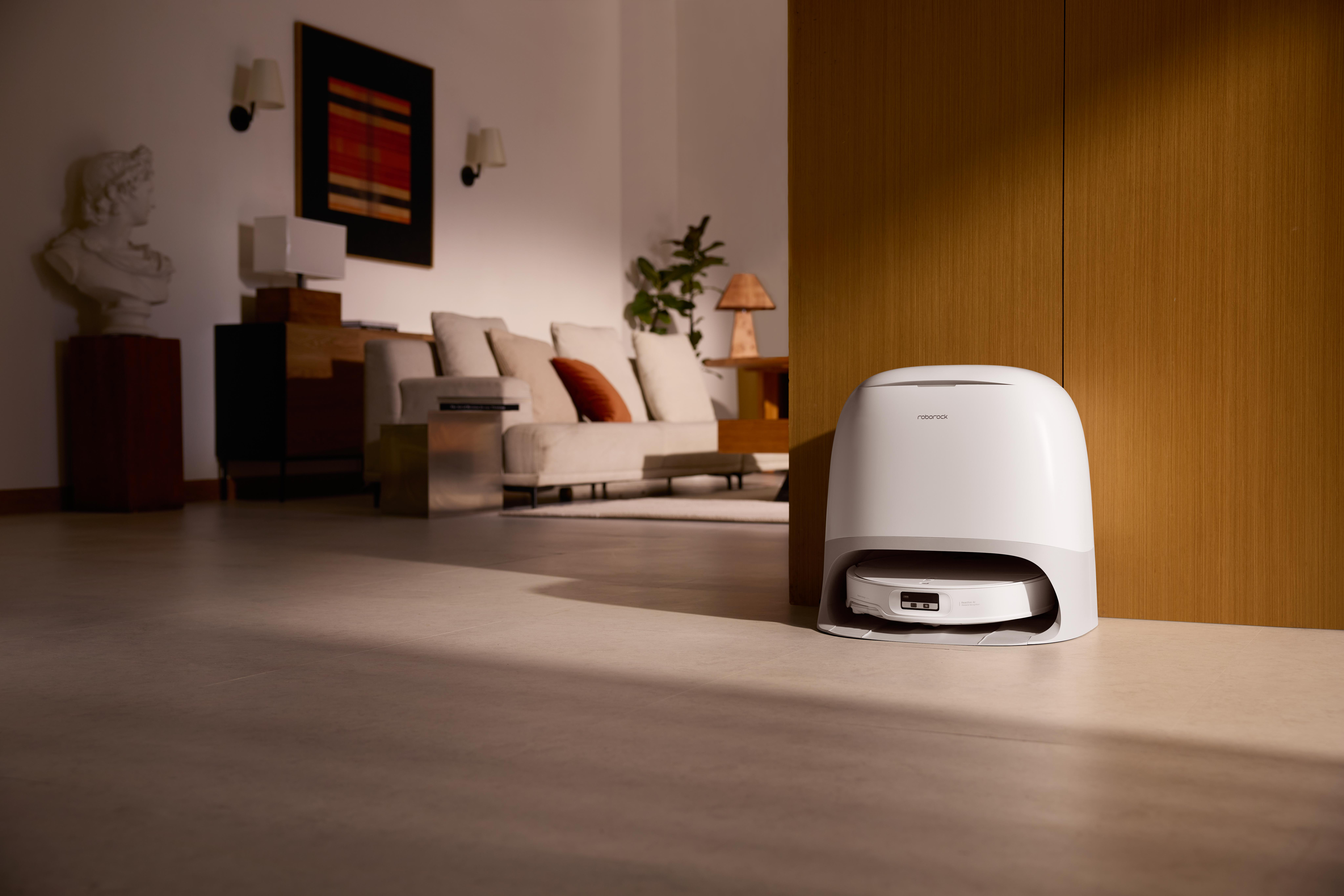 Ein weißer Roboter-Staubsauger steht neben einer Holzwand. Im verschwommenen Hintergrund sind ein Sofa und Kunst zu sehen.
