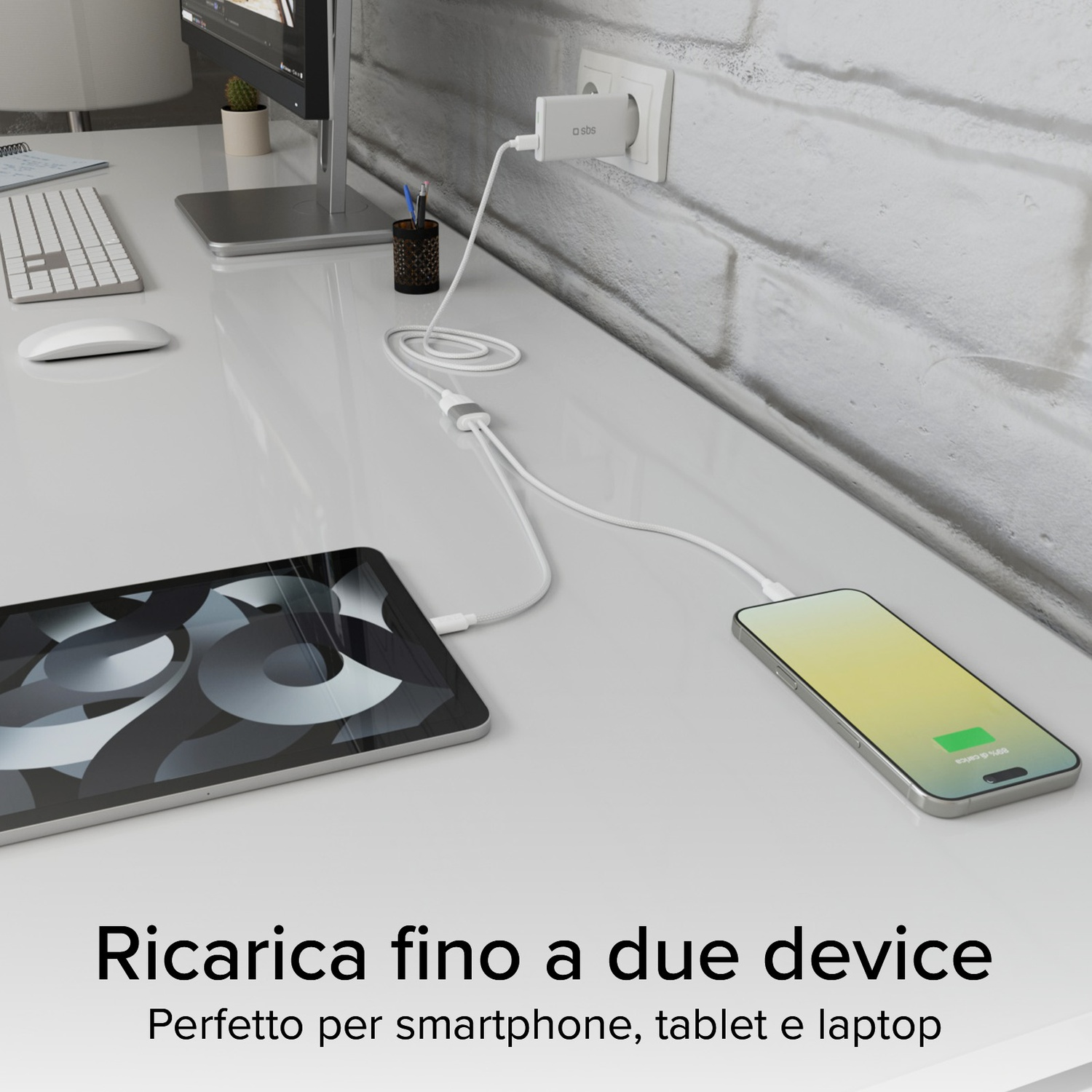 Scrivania bianca con telefono e tablet in carica. Caricatore collegato al muro. Monitor, tastiera e mouse visibili.