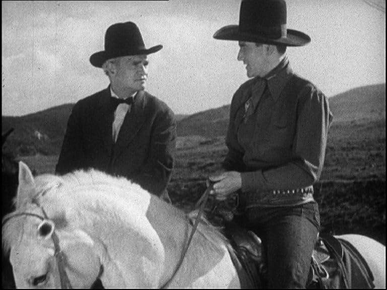 Zwei Männer mit Hüten auf Pferden, einer im Anzug, der andere im Cowboy-Outfit, unterhalten sich in einer Landschaft.