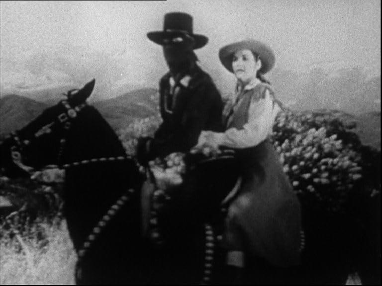 Ein maskierter Mann und eine Frau auf einem Pferd. Berge sind im Hintergrund. Die Szene ist in Schwarzweiß.