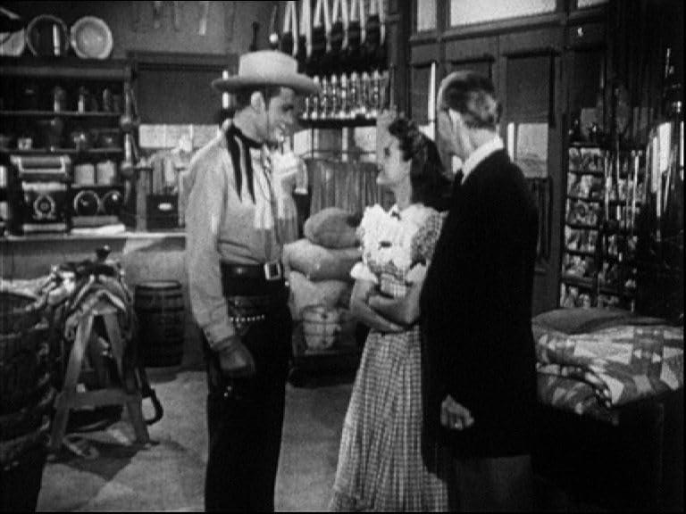 Ein Cowboy mit einer Frau und einem Mann in einem Geschäft. Der Cowboy trägt einen Hut und ein Halstuch. Die Frau trägt ein Kleid. Der Mann trägt einen Anzug.