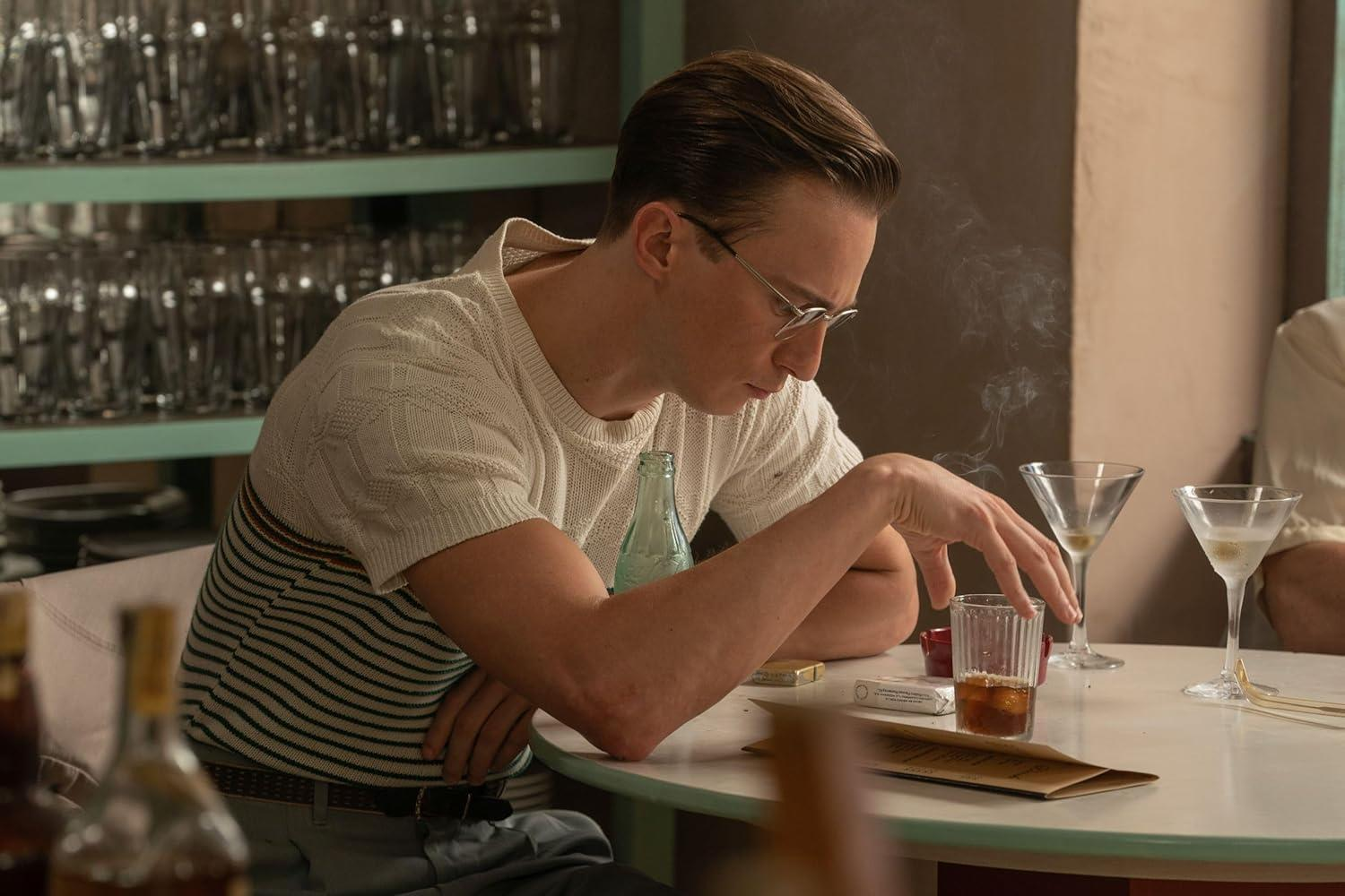 Ein Mann mit Brille sitzt am Tisch und raucht. Auf dem Tisch stehen Getränke.