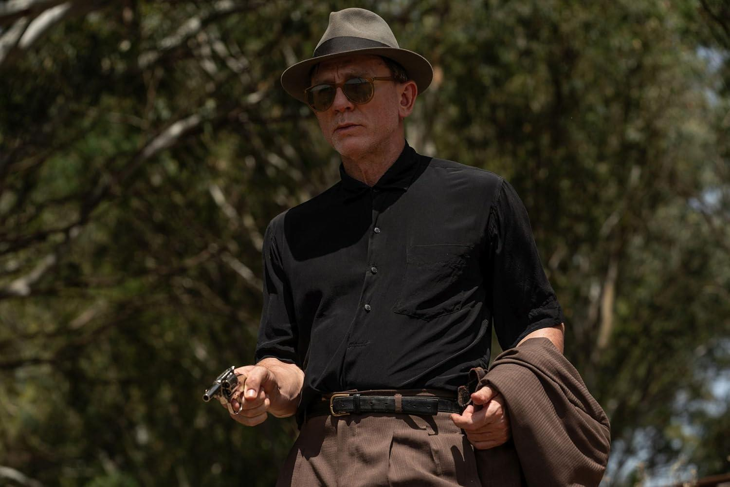Ein Mann mit Hut und Sonnenbrille hält eine Pistole und eine Jacke in einer baumbestandenen Umgebung.
