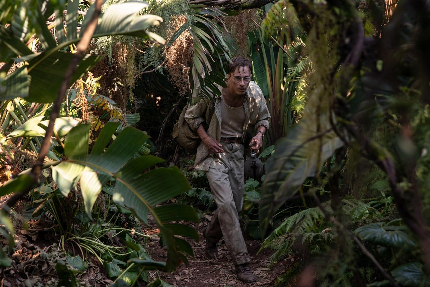 Ein Mann in abgetragener Kleidung mit Rucksack wandert durch einen dichten, grünen Dschungel.