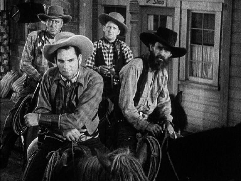 Vier Cowboys auf Pferden vor einem Laden. Sie tragen Hüte und sehen streng aus. Schwarz und Weiß.