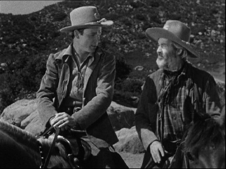 Zwei Männer mit Cowboyhüten sitzen auf Pferden. Einer trägt eine Jacke, der andere ein kariertes Hemd. Felsiger Hintergrund.
