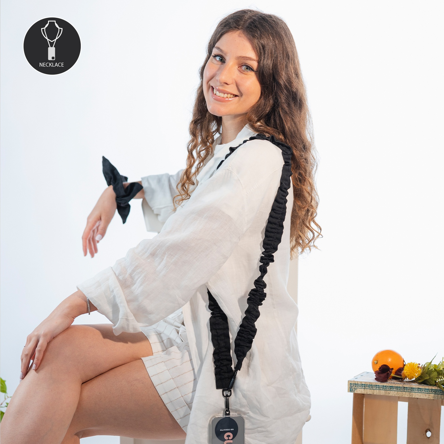 Donna in camicia bianca sorride, indossa un cordino nero. Arancia e fiori su un tavolino di legno.