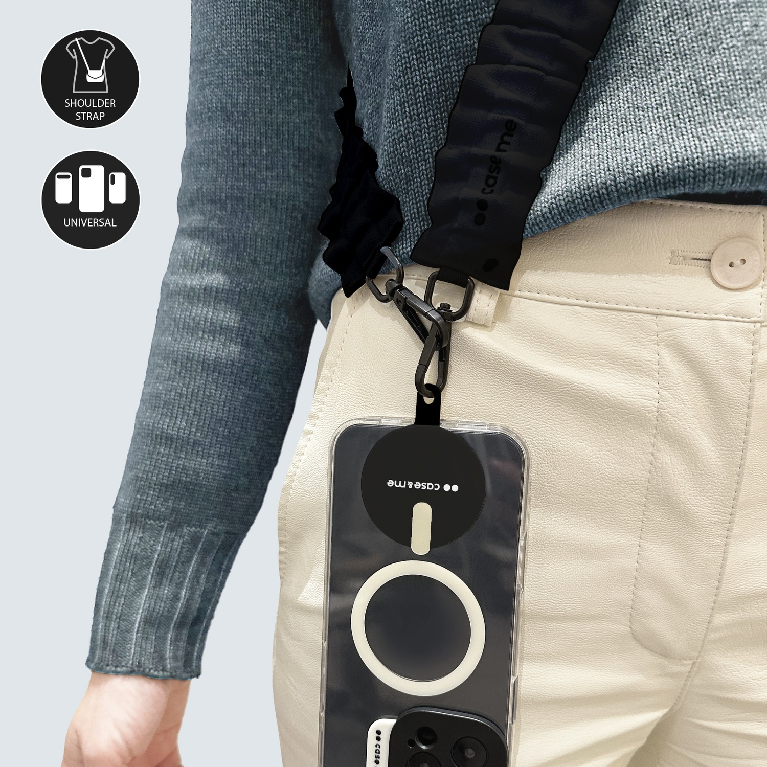 Persona con maglione e pantaloni color crema, tiene in mano un telefono con cinturino nero. Il telefono ha un cerchio bianco e una custodia nera.