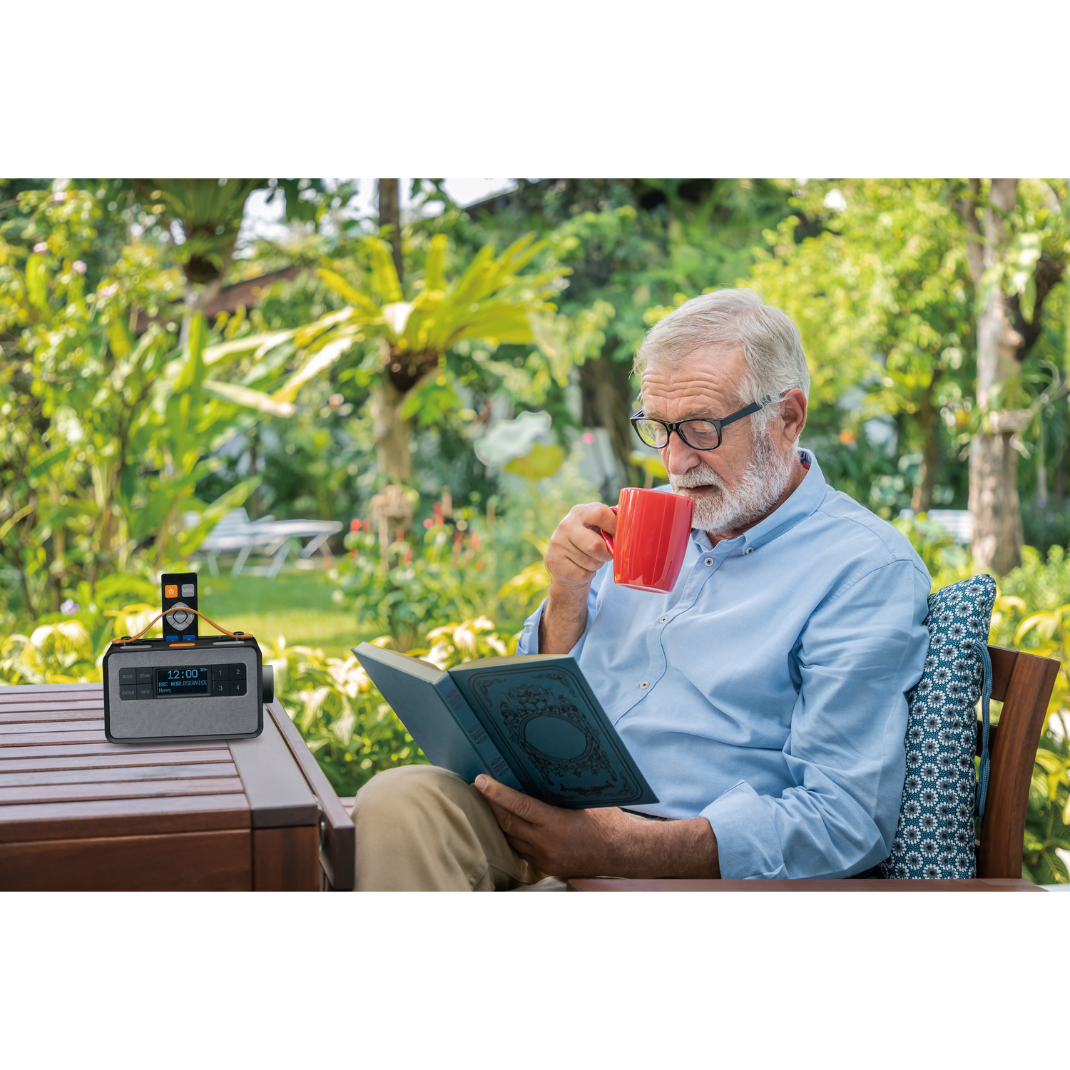 Homme âgé avec des lunettes boit d'une tasse rouge, lisant un livre en plein air, radio sur la table, jardin en arrière-plan.