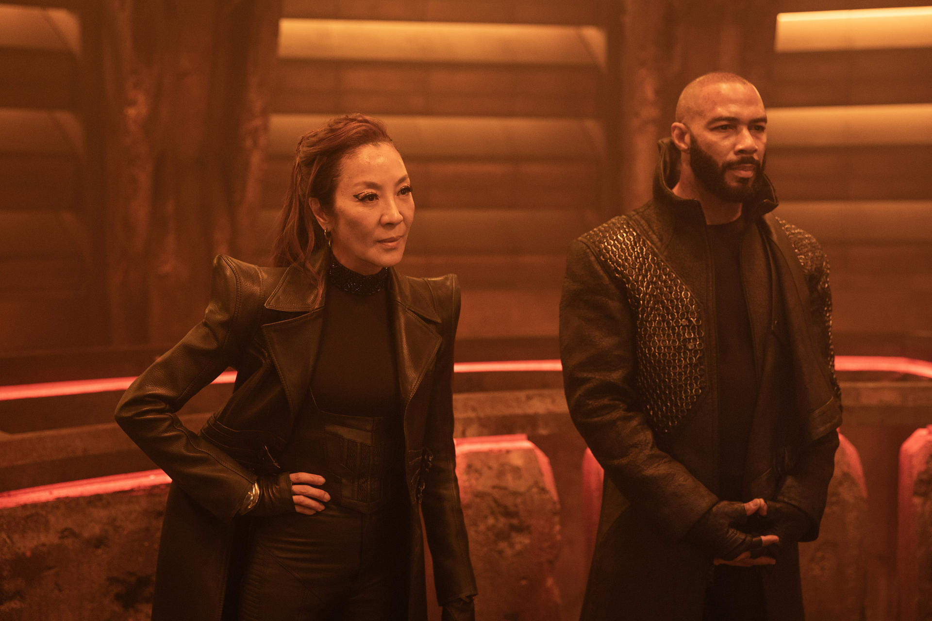 Zwei Personen in schwarzen Outfits stehen in einem schwach beleuchteten Raum. Eine Frau hat rotes Haar. Ein Mann mit Bart steht neben ihr.