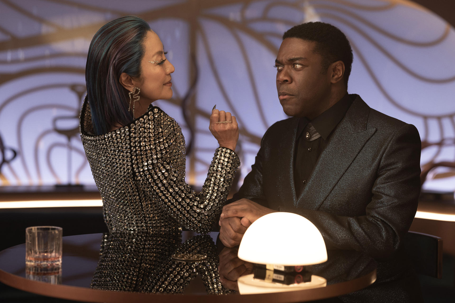 Zwei Personen an einem Tisch. Eine Frau in einem mit Juwelen besetzten Oberteil blickt einen Mann im Anzug an. Sie scheinen sich zu unterhalten.