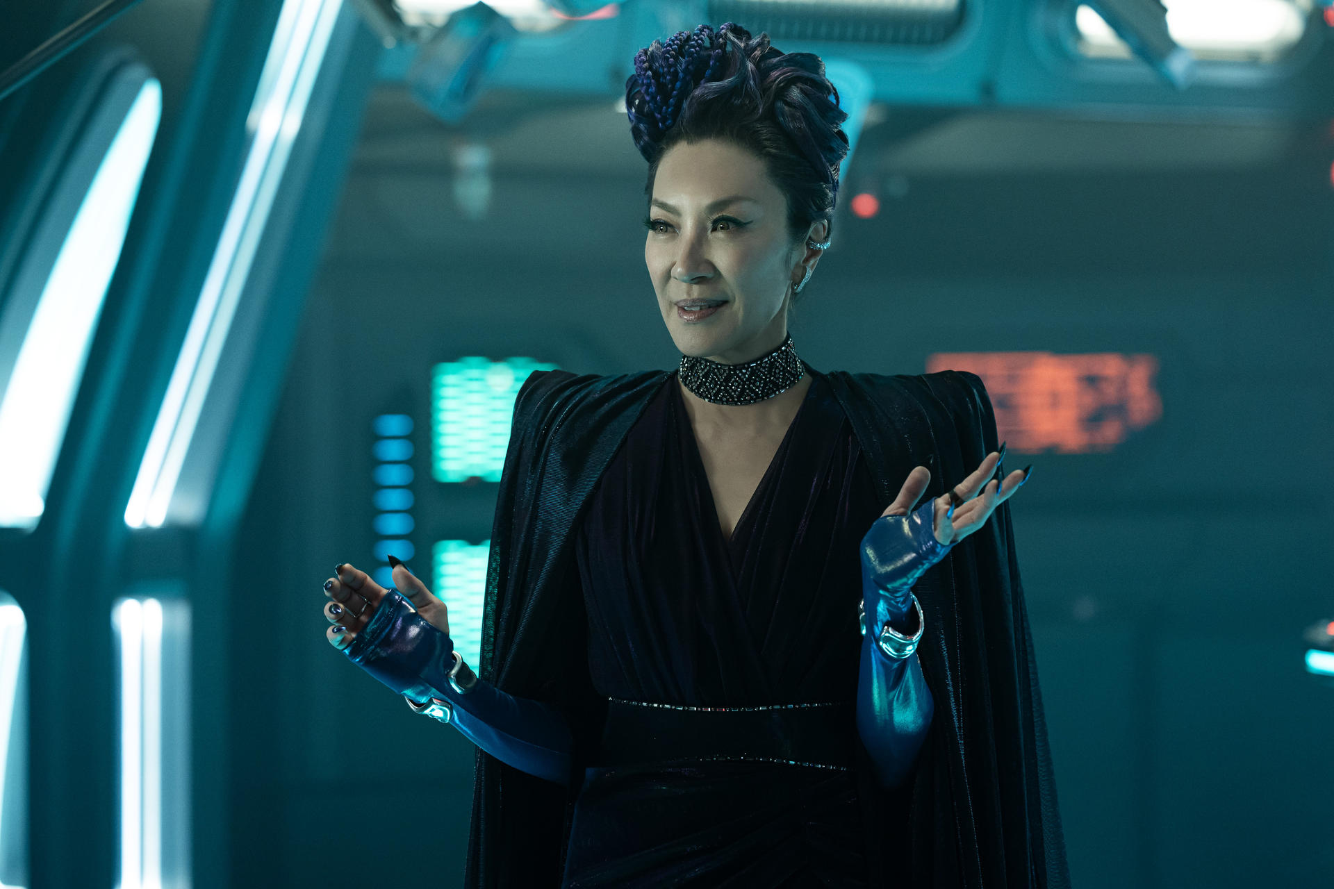 Frau mit lila Haaren in dunkler Kleidung gestikuliert in futuristischer Umgebung. Hintergrund hat blaue Lichter und Paneele.