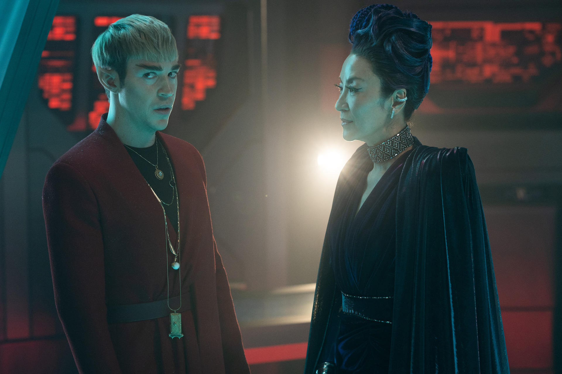 Zwei Personen in futuristischer Kleidung stehen sich gegenüber. Einer trägt Rot, der andere Dunkelblau. Der Hintergrund ist ein dunkler, technischer Raum.