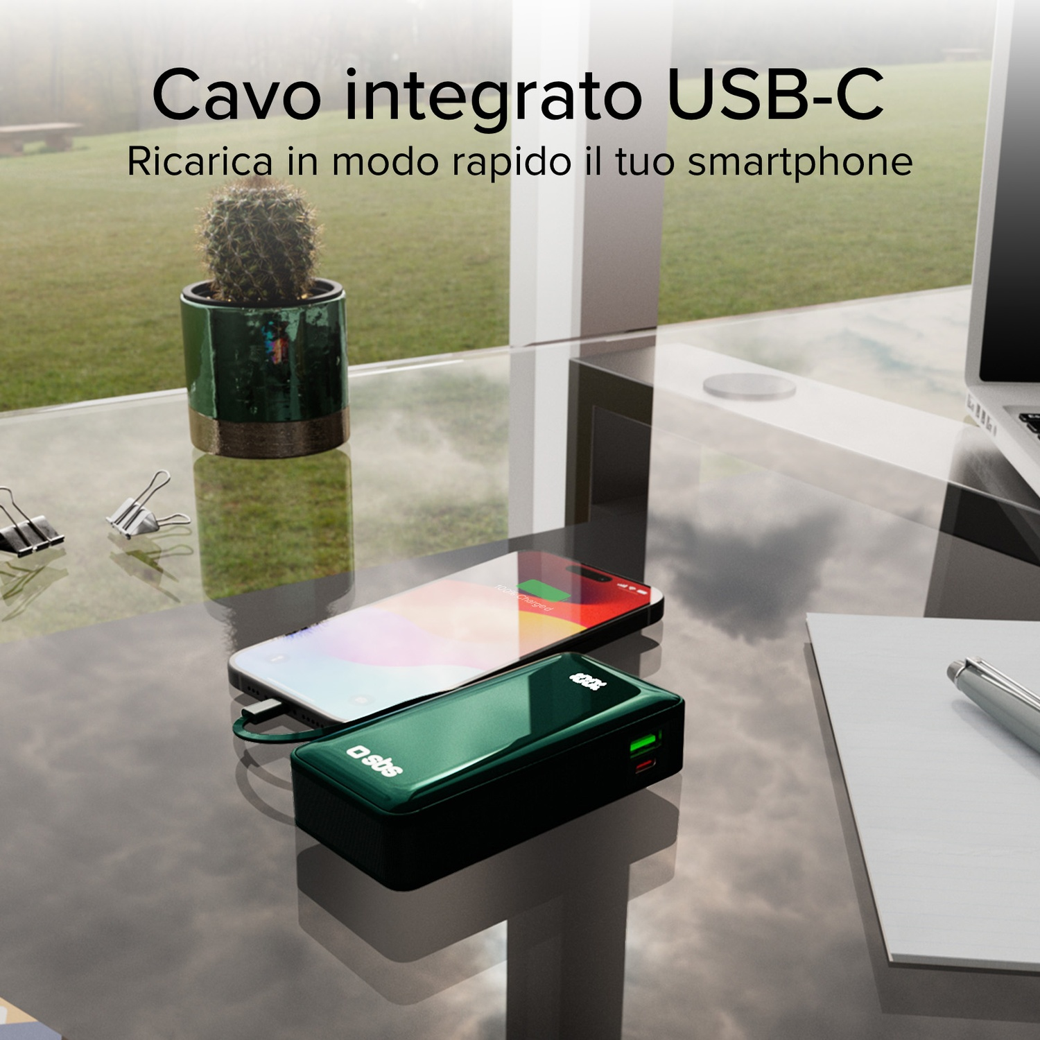 Un power bank verde carica un telefono. Un cactus in vaso e un laptop sono anche sul tavolo di vetro.