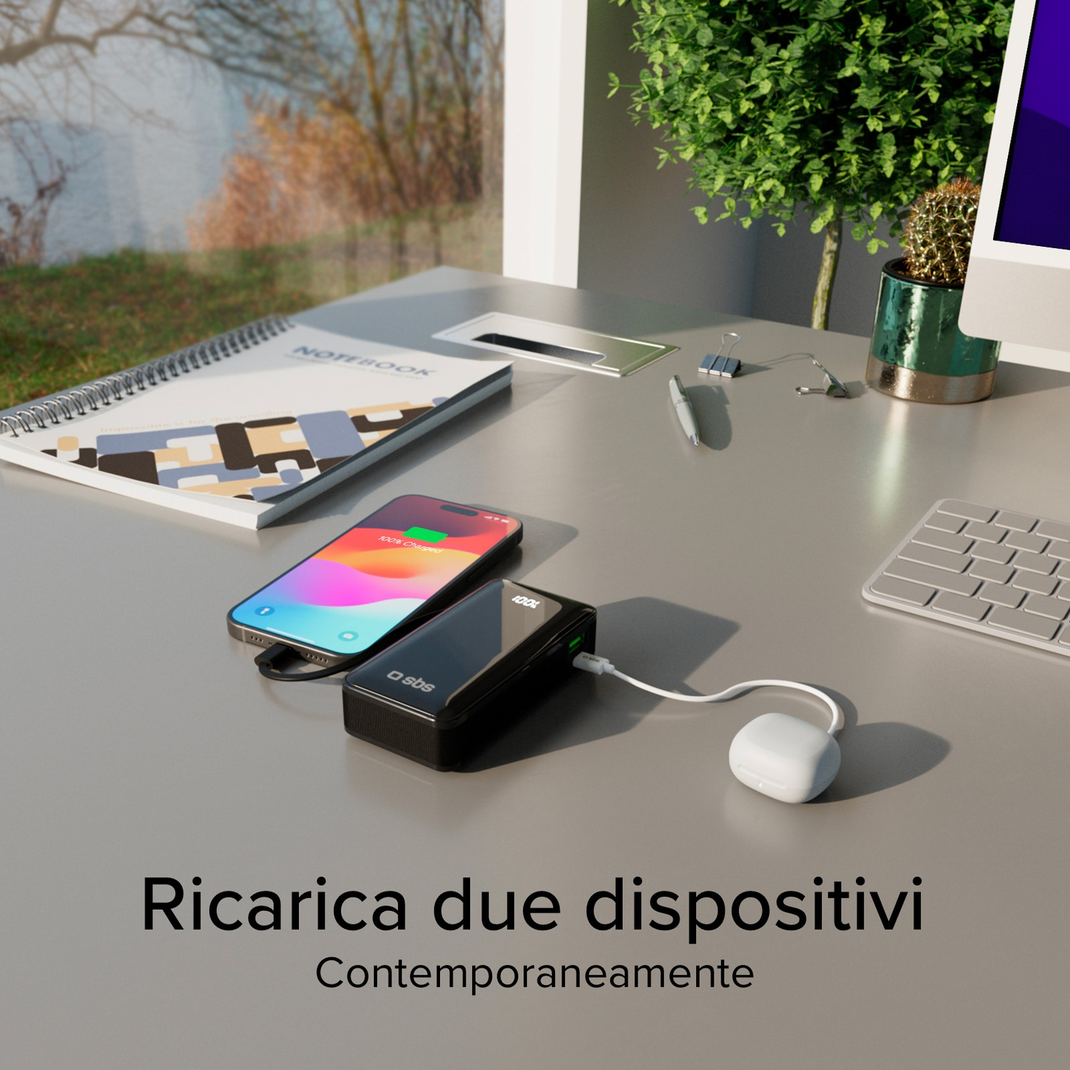 Scrivania con telefono in carica, quaderno, piccola pianta e finestra con vista. Il telefono è carico al 100%.