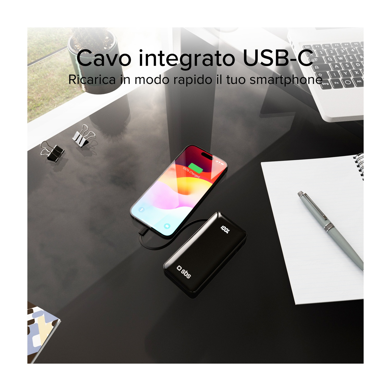 Un telefono in carica tramite un power bank nero. Un taccuino, una penna e un laptop sono anche sulla scrivania nera.