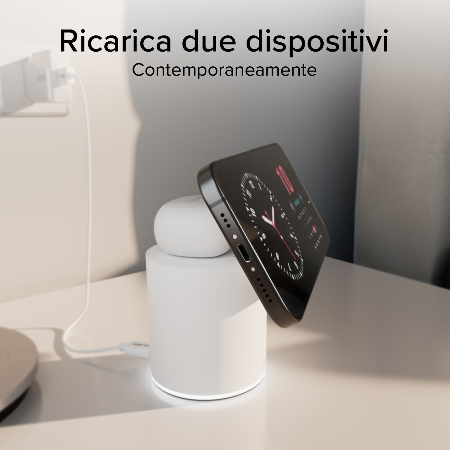 Un telefono e auricolari in carica su una base bianca. Caricatore collegato al muro. Testo: Ricarica due dispositivi contemporaneamente.