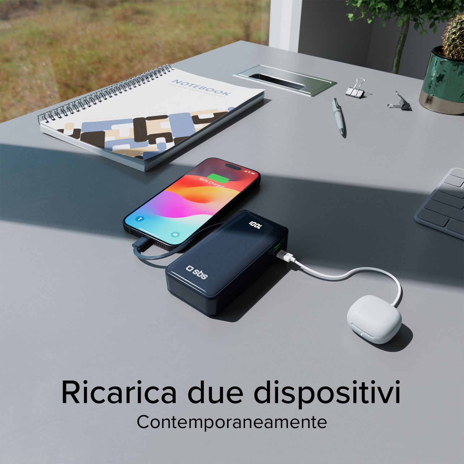 Un telefono e un power bank in carica su una scrivania. Un quaderno, una penna, graffette e una pianta sono anche sulla scrivania.