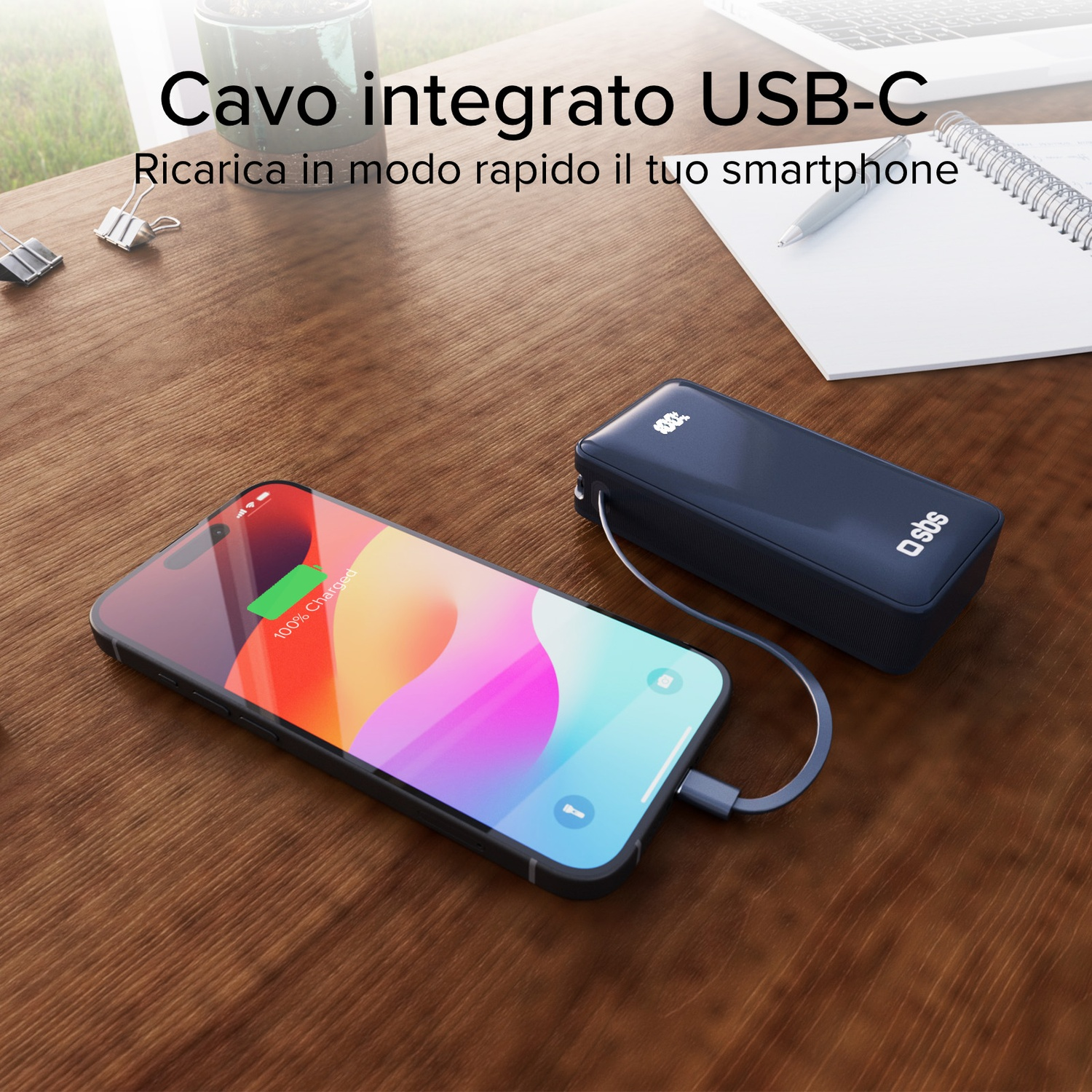 Uno smartphone si carica da un power bank blu scuro tramite un cavo USB-C su una superficie di legno.