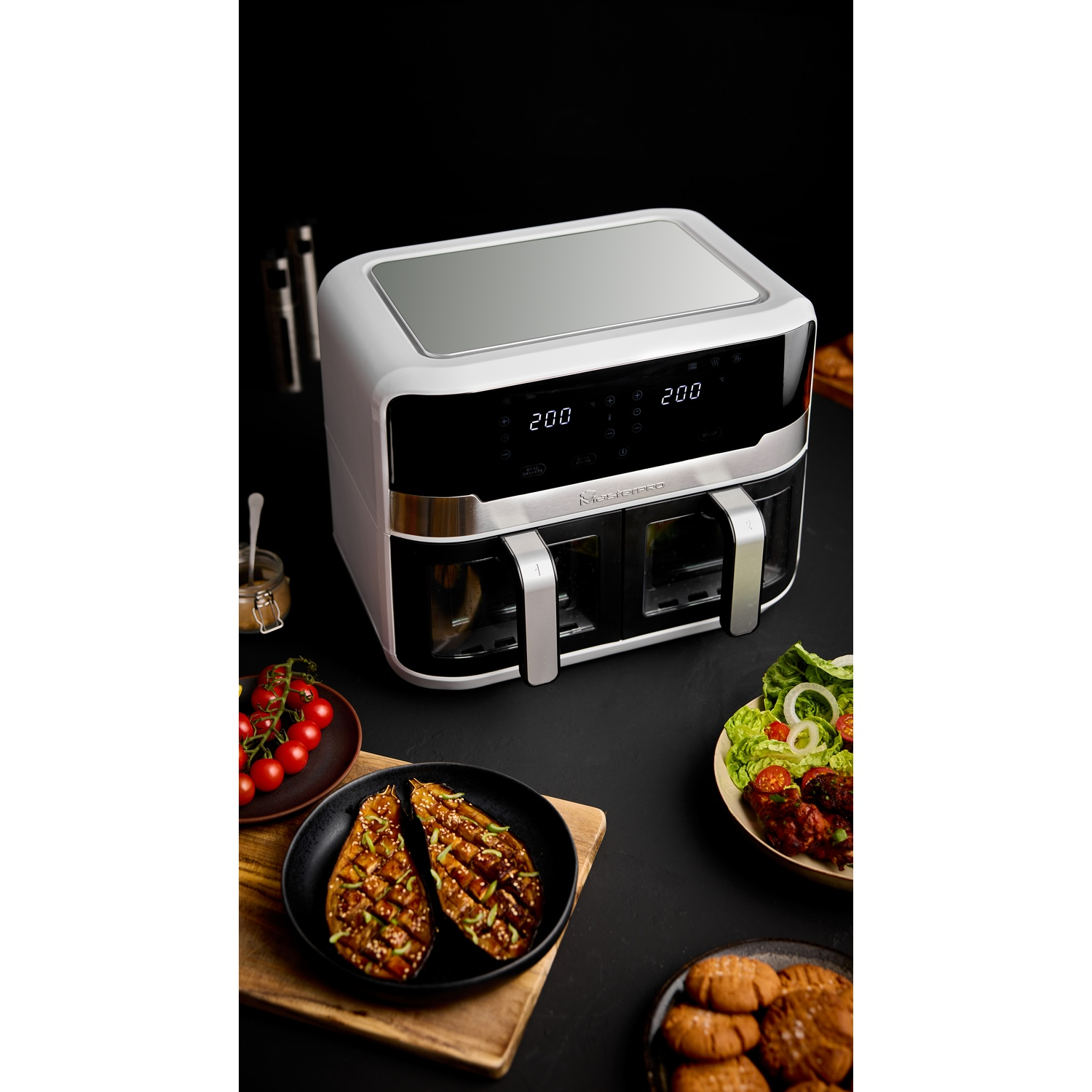 Friggitrice ad aria bianca con cibo su una superficie nera. Include melanzane grigliate, pomodori, insalata, biscotti e altri piatti.