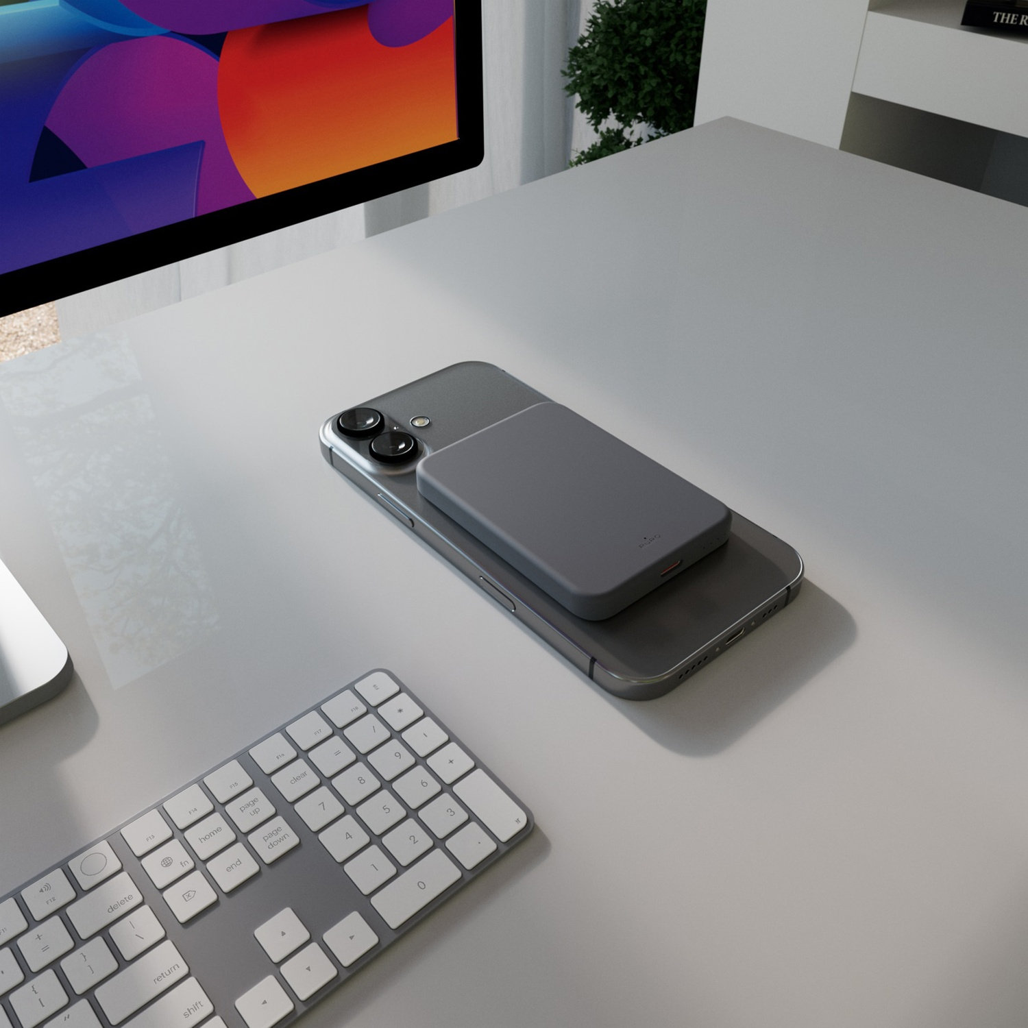 Un telefono grigio con un caricabatterie grigio è su una scrivania bianca. Sono visibili una tastiera e un monitor con display colorato.