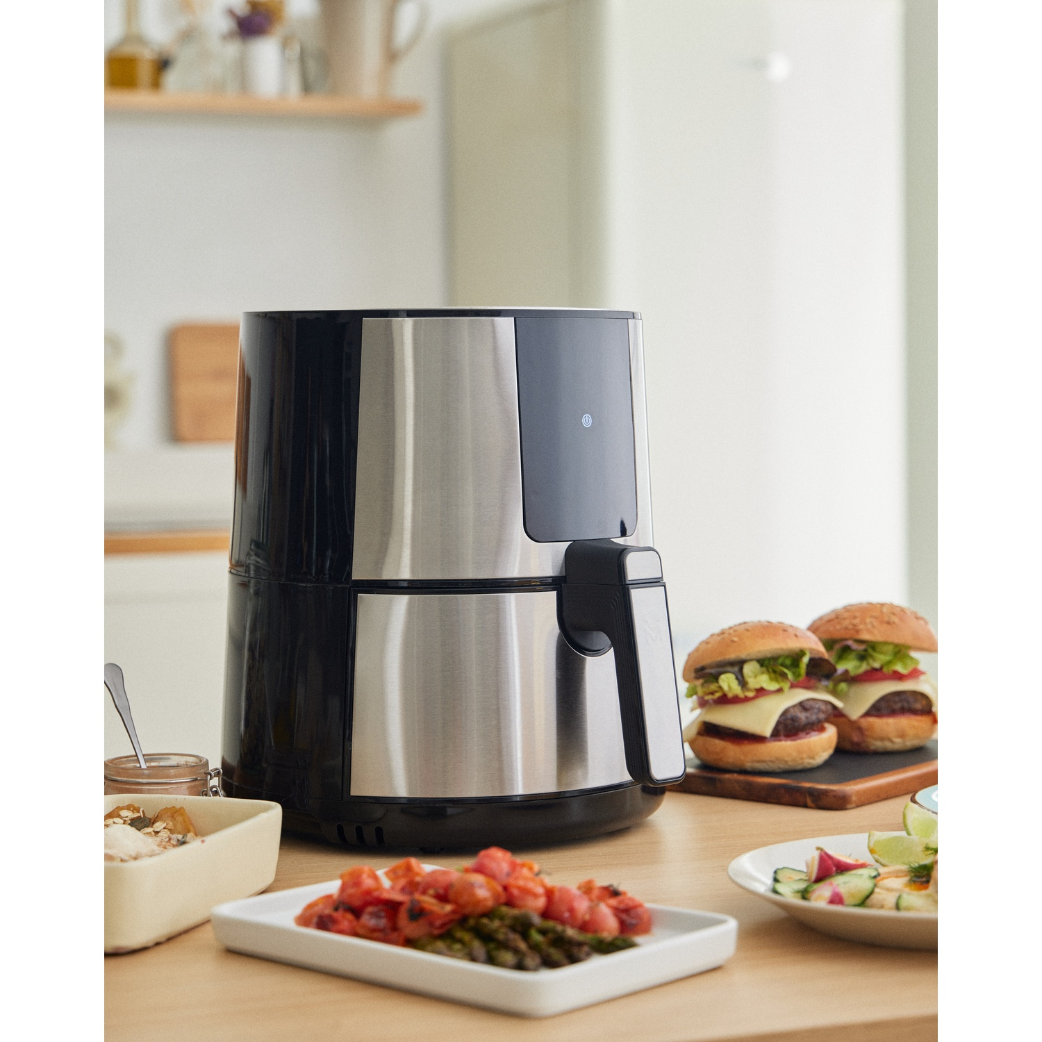 Friggitrice ad aria con hamburger, insalata e pomodori su un tavolo. Ambiente cucina con frigorifero bianco sullo sfondo.
