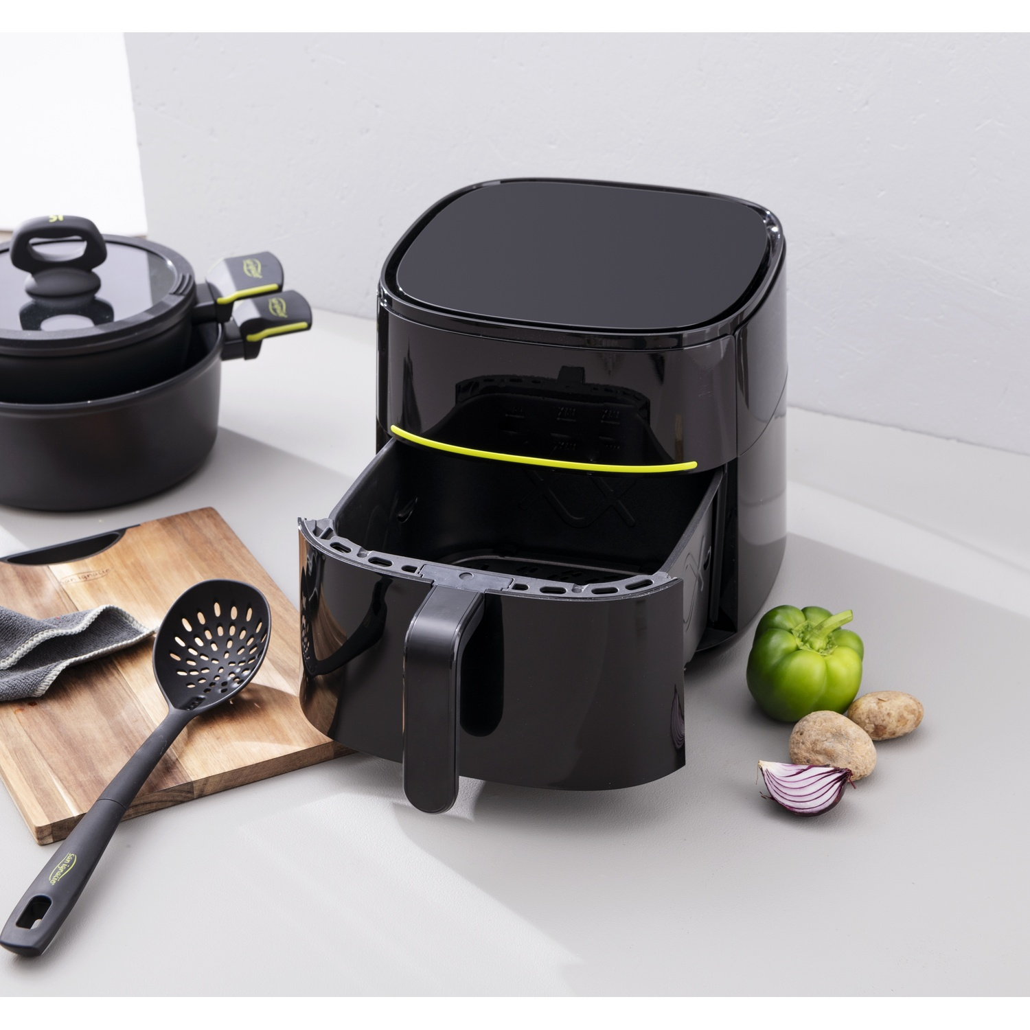 Friggitrice ad aria nera con cassetto aperto, pentola, tagliere, cucchiaio, verdure sul piano di lavoro. Sfondo grigio.