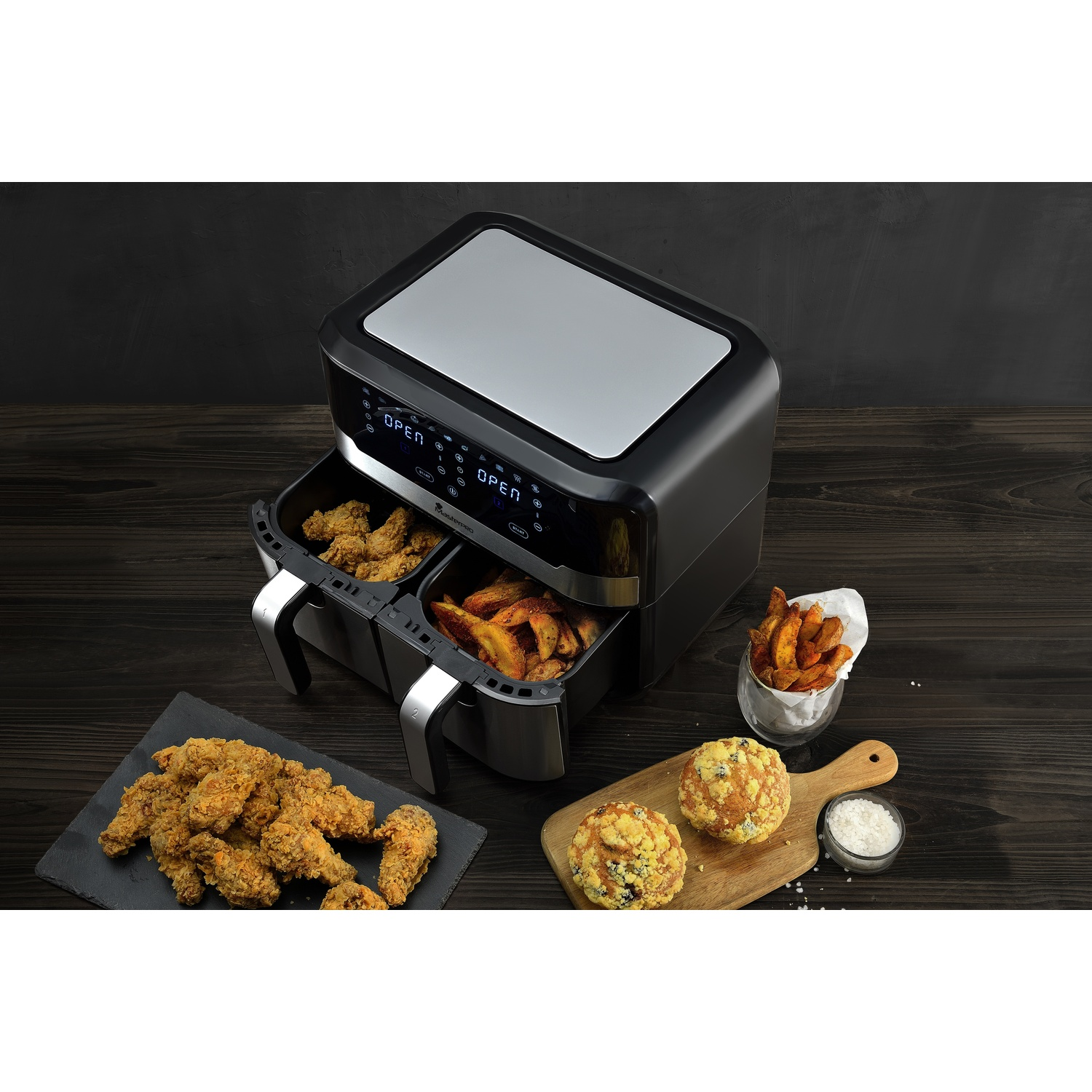 Una friggitrice ad aria con pollo fritto, patatine fritte e due muffin su un tavolo di legno. Lo sfondo è un muro scuro.