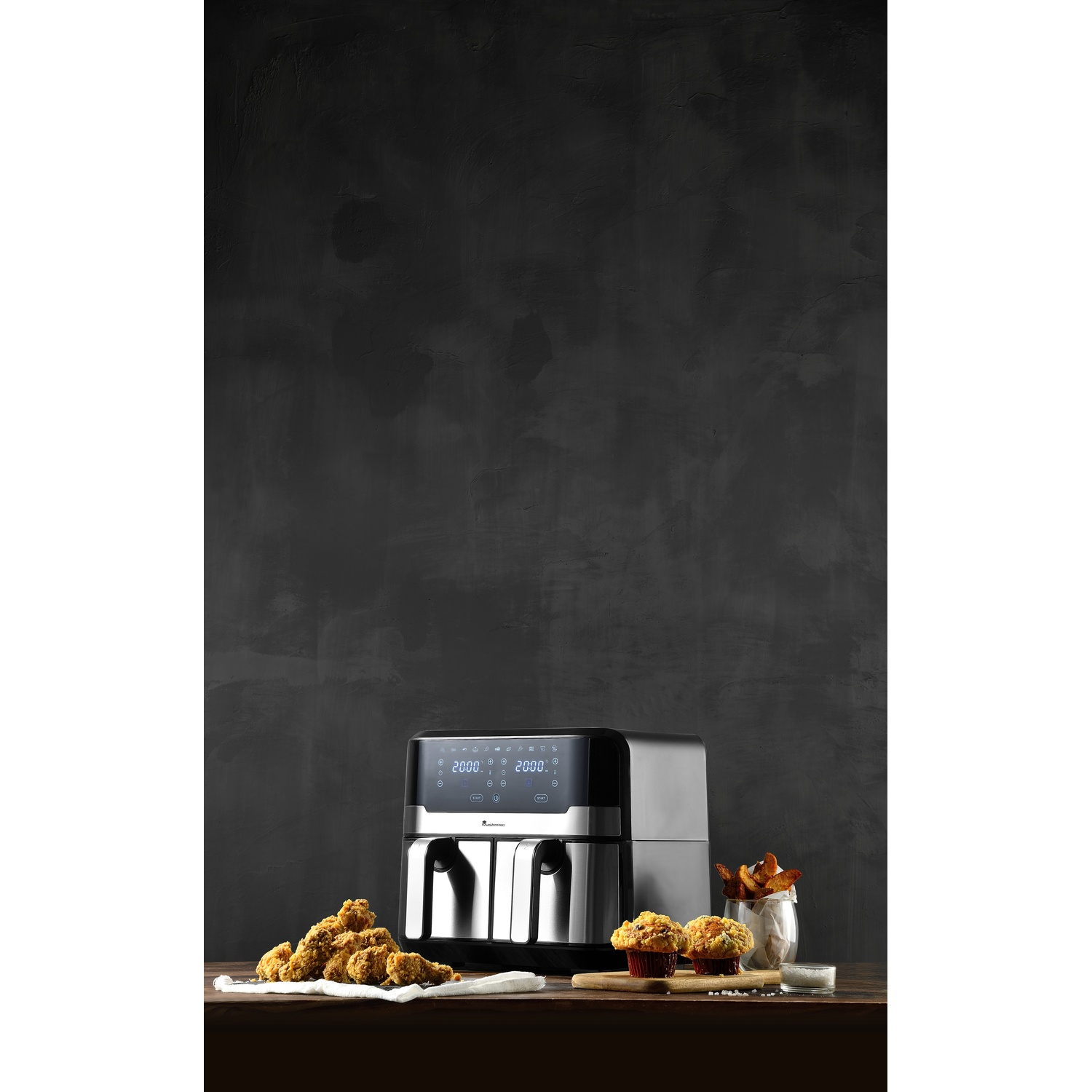 Una friggitrice ad aria, pollo fritto, muffin e patatine fritte su un tavolo. Sfondo grigio scuro.