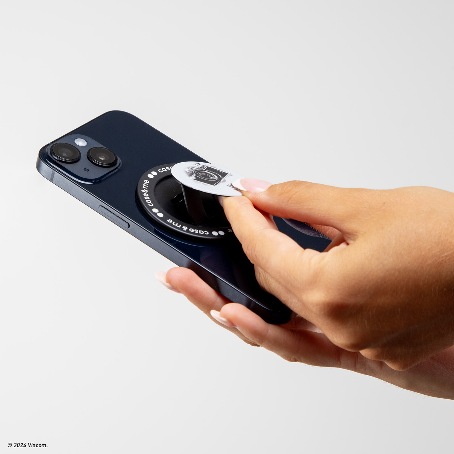 Mano che tiene un telefono con un'impugnatura per fotocamera. Il telefono è blu scuro e l'impugnatura ha un'immagine della fotocamera. Sfondo bianco.