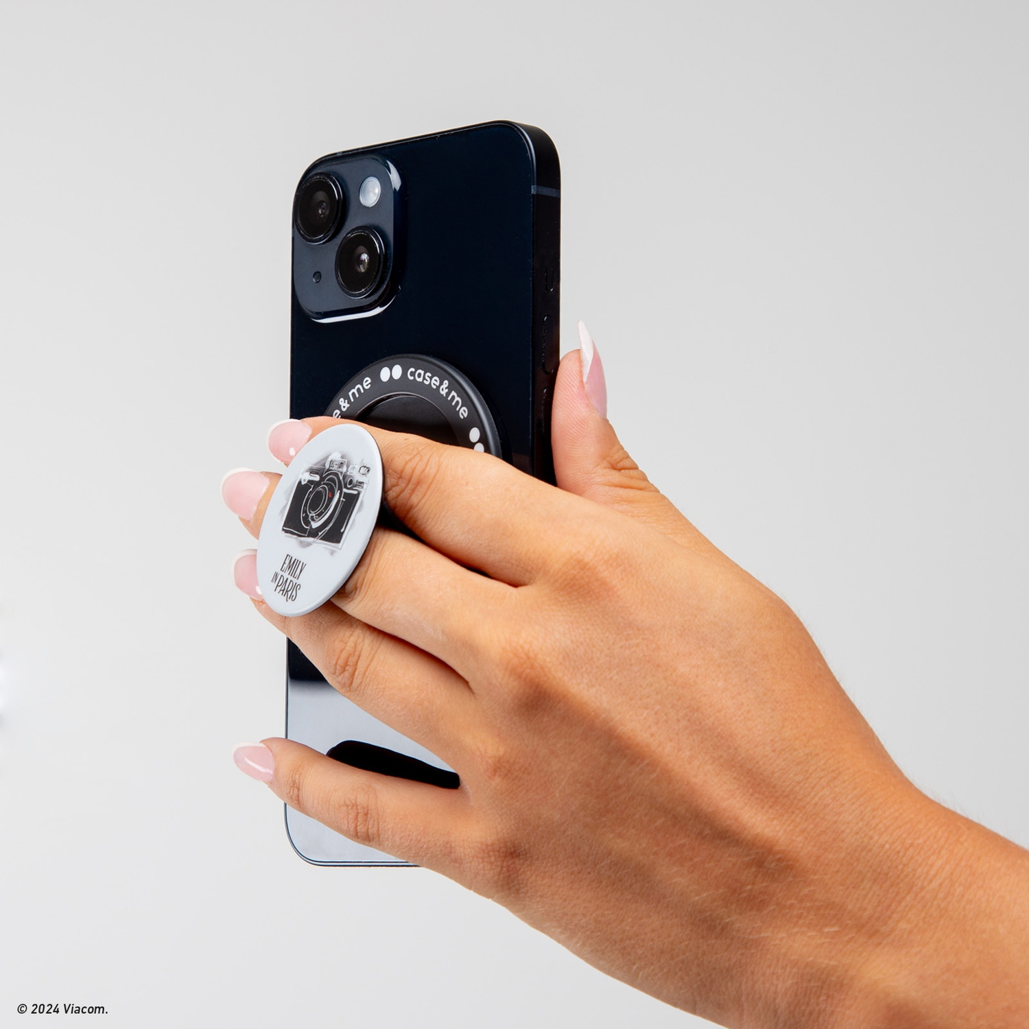 Una mano tiene un telefono nero con un pop socket a tema fotocamera. Lo sfondo è bianco.