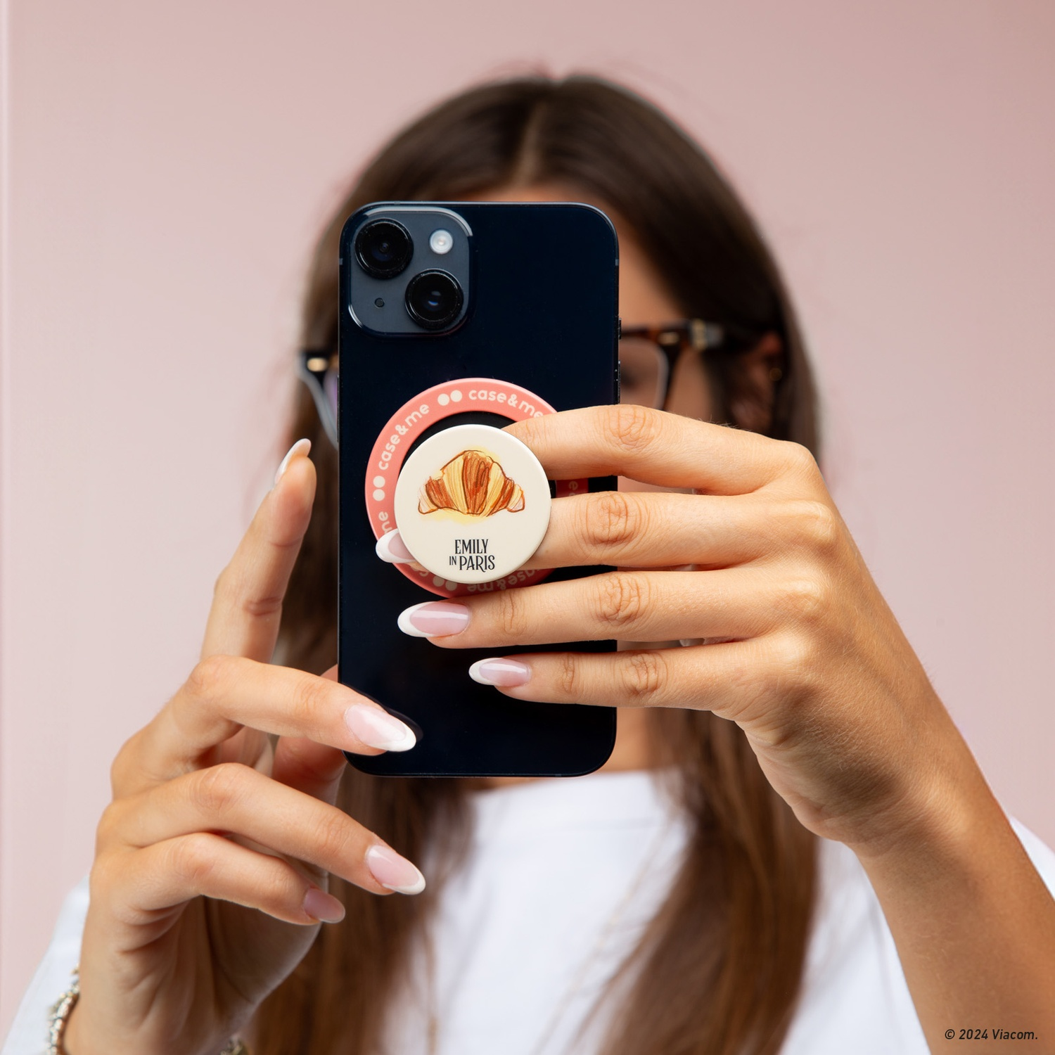 Una donna tiene un telefono nero con un pop socket che dice "Emily in Paris" e mostra un croissant, su sfondo rosa.