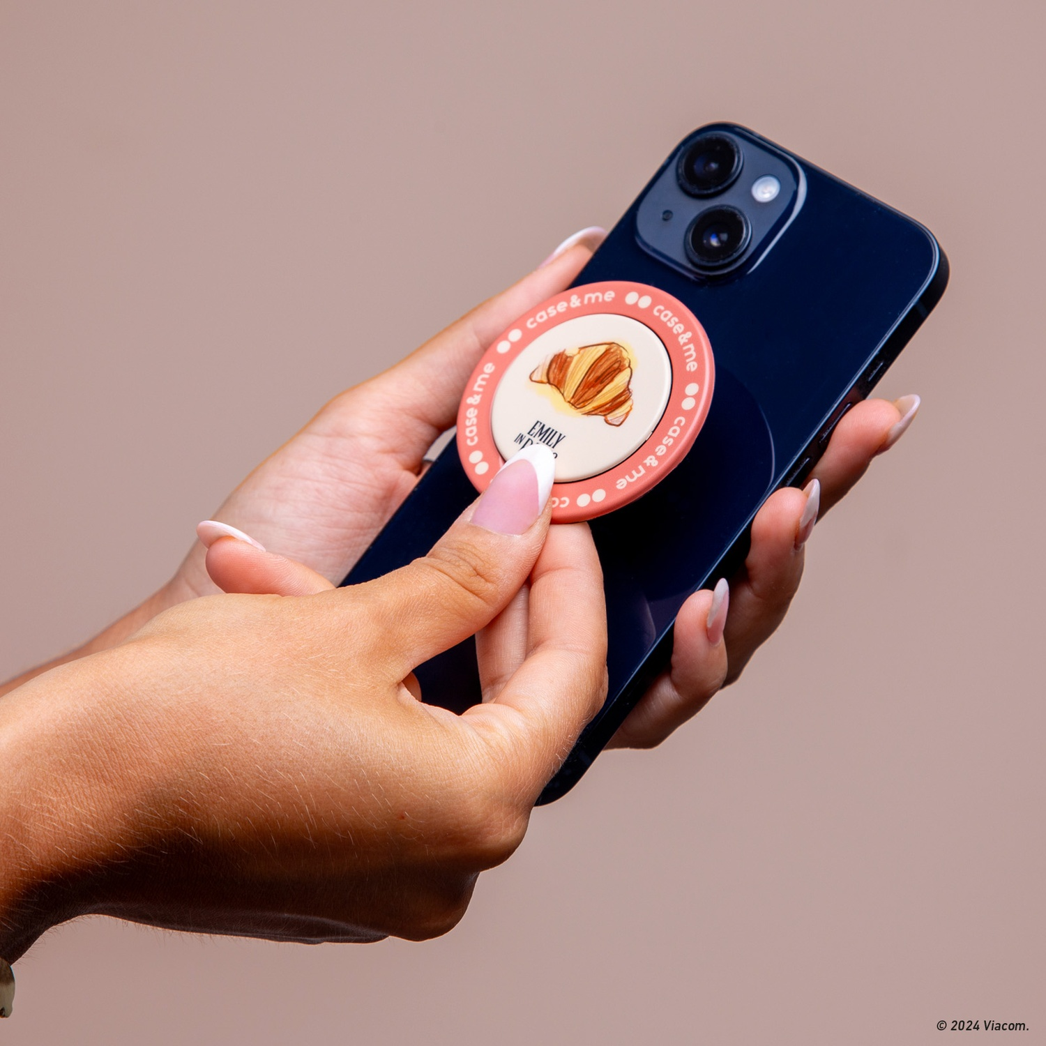 Mani che tengono un telefono blu scuro con un pop socket. Il pop socket ha un'immagine di croissant e la scritta "case & me".