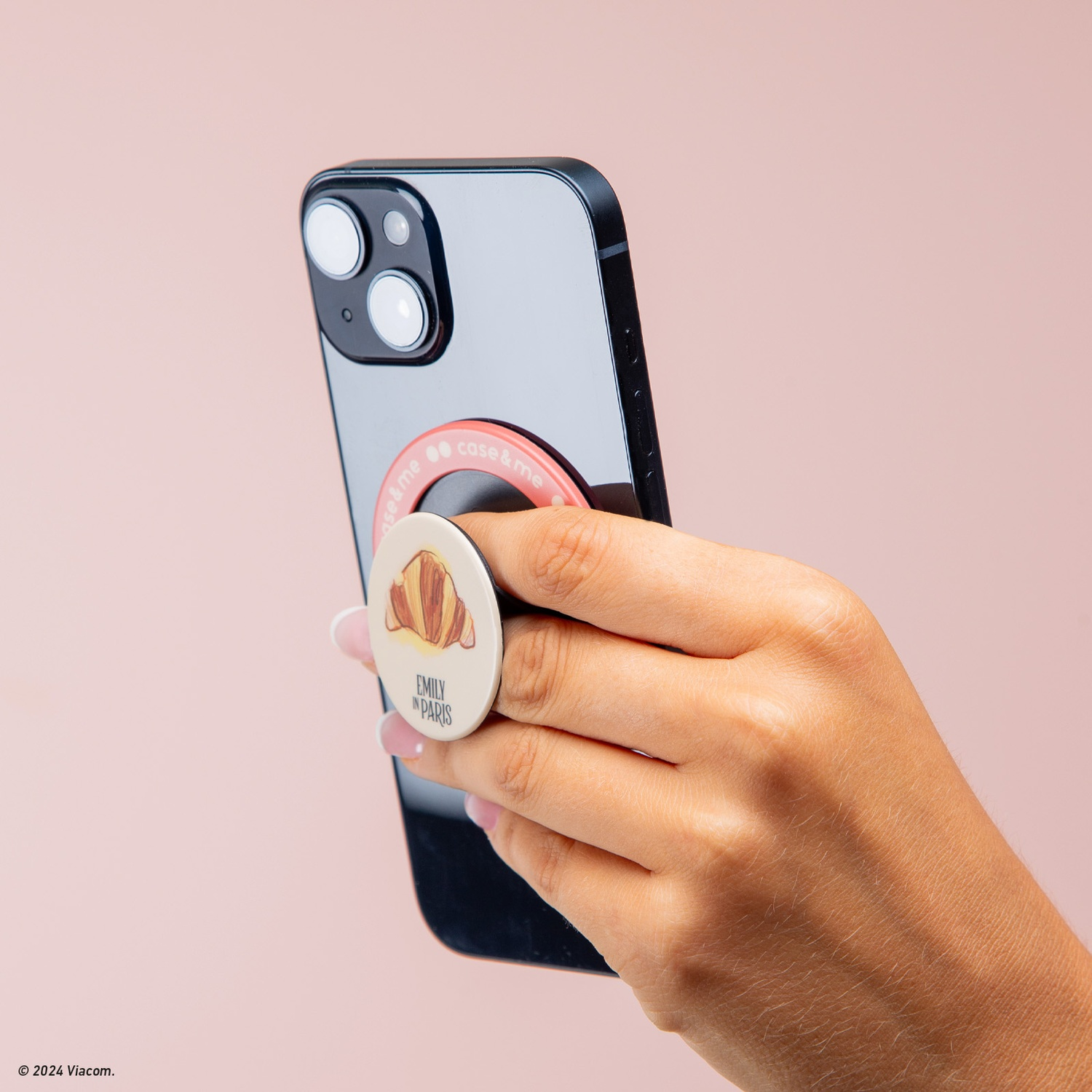 Una mano tiene un telefono nero con un popsocket con un croissant e "Emily in Paris" su sfondo rosa.