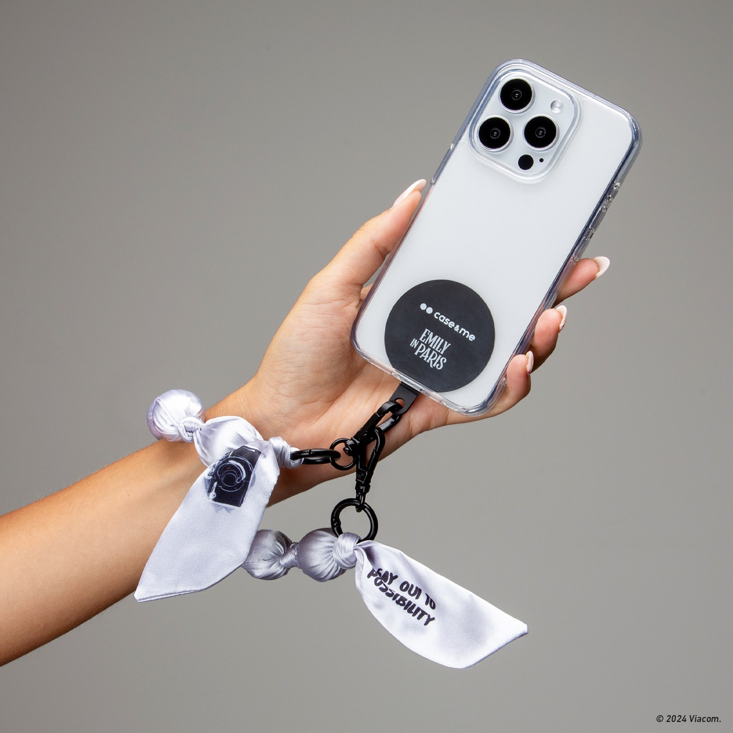 Una mano tiene un telefono in una custodia trasparente. Attaccato c'è un braccialetto grigio chiaro con una fotocamera e le parole "Say Oui to Possibility".