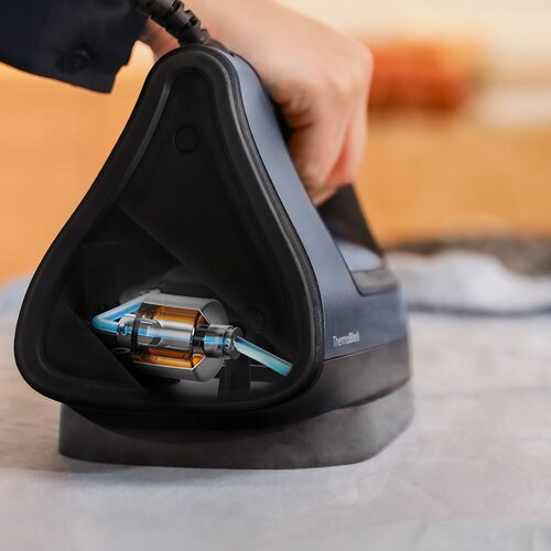 Ręka trzyma niebiesko-czarne żelazko. W środku widoczny jest szczegółowy mechanizm. Żelazko leży na białej tkaninie.